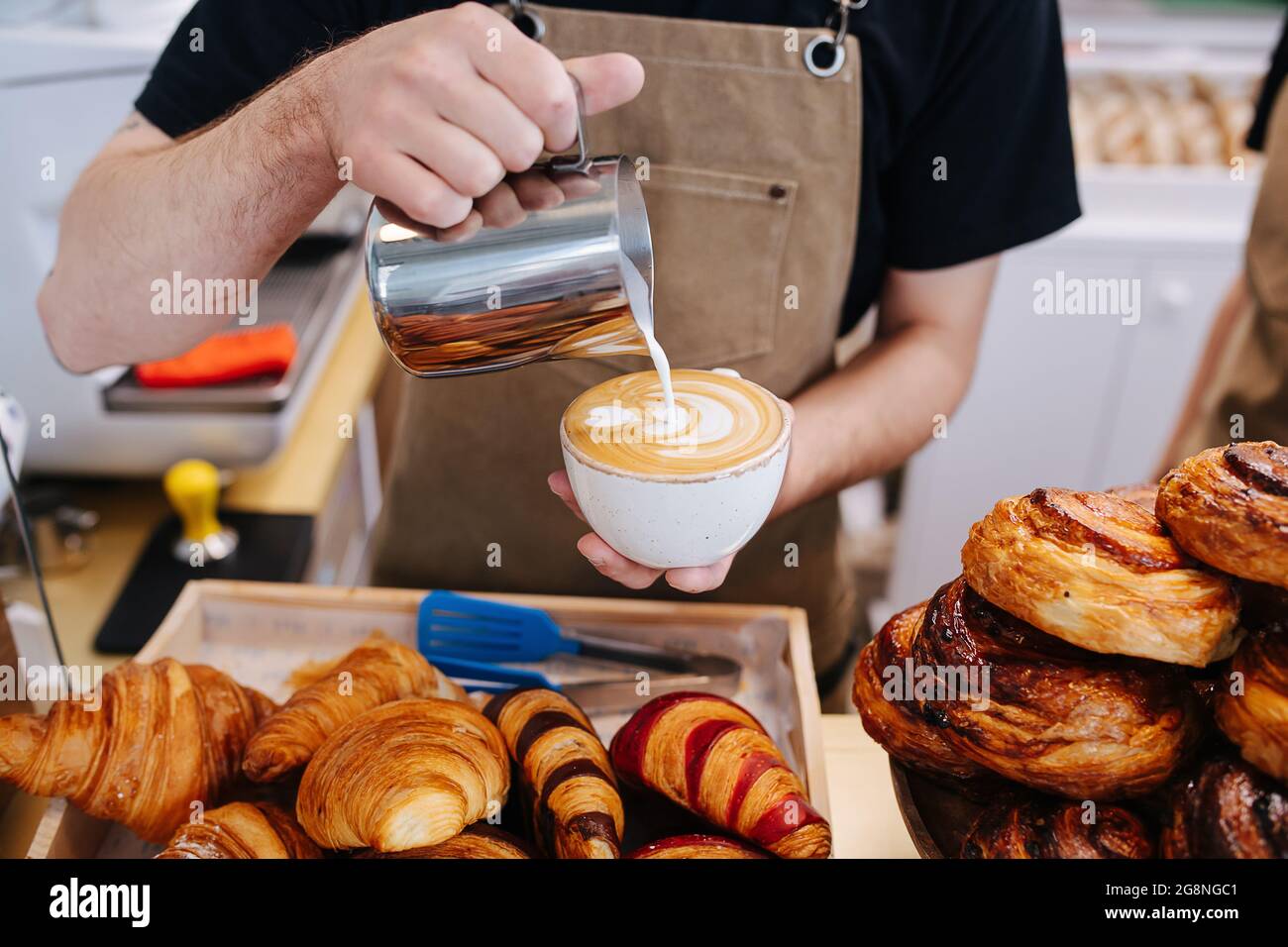 Image rognée d'un boulanger masculin mettant de la crème dans une tasse de café d'une verseuse métallique. Dans une cuisine de boulangerie à côté d'une pile de pâtisserie. Banque D'Images