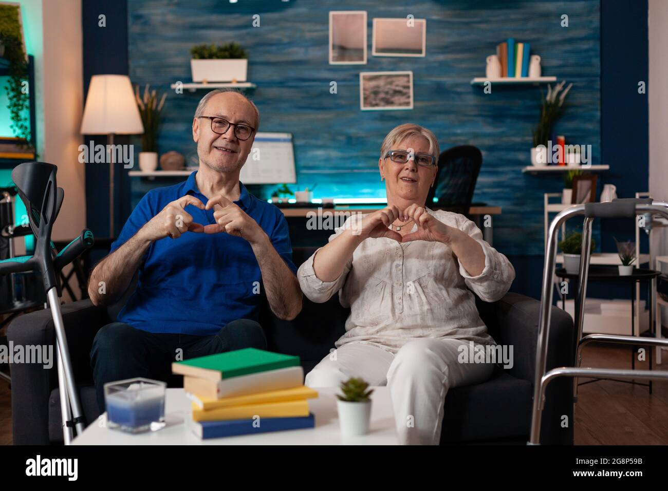 Vieux caucasien heureux couple faisant coeur signe avec les mains tout en regardant la caméra gaie assis dans la salle de séjour à la maison. Homme et femme gaie à la retraite montrant le symbole de l'amour romantique Banque D'Images