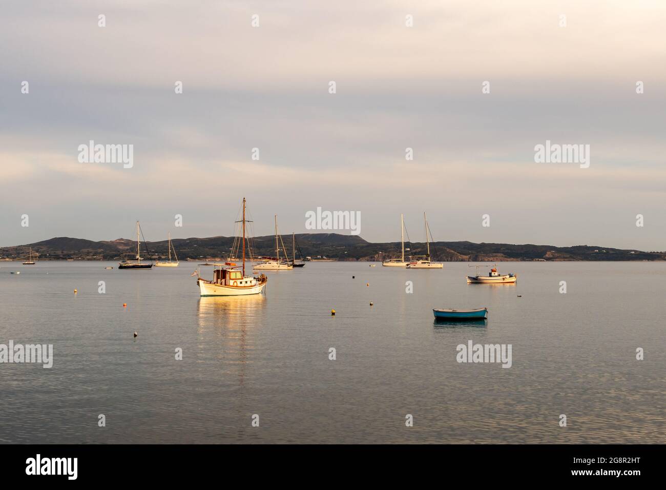Bateaux de pêche et de voile à mouillage dans le port d'Adamas sur l'île de Milos, Grèce pendant l'heure d'or, avec mer calme plate et ciel nuageux. Banque D'Images