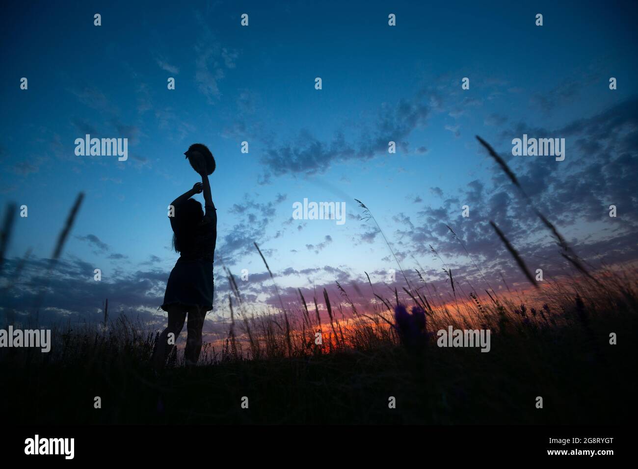 Femme solitaire marchant sur le champ d'herbe sur le fond de crépuscule d'été, les mains soulevées au ciel et tenant chapeau de paille Banque D'Images