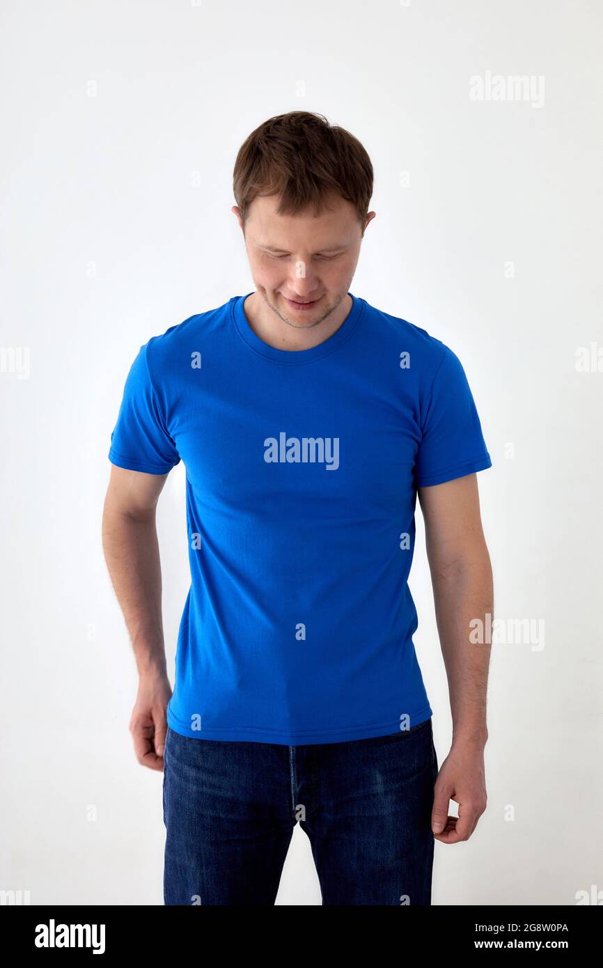 Jeune homme en Jean avec main derrière et t-shirt bleu debout sur fond blanc et regardant vers le bas Banque D'Images