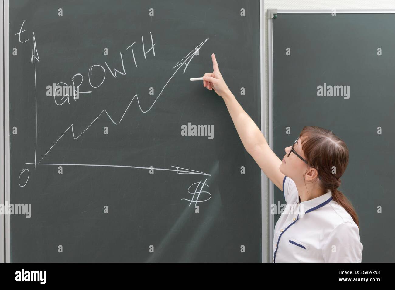 une jeune femme entraîneur d'affaires dans un chemisier blanc et des lunettes au tableau noir dans le bureau dessine un graphique de profit. gros plan Banque D'Images