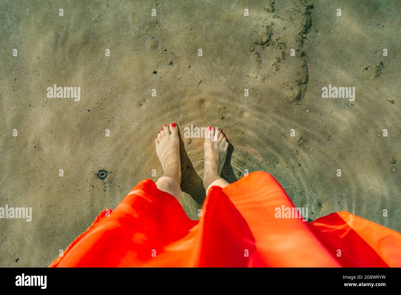 Bonne femme en robe rouge debout sur la plage, appréciant la journée ensoleillée d'été, les pieds sont couverts de sable et de l'eau de mer cristalline.femmes jambes pieds nus Banque D'Images