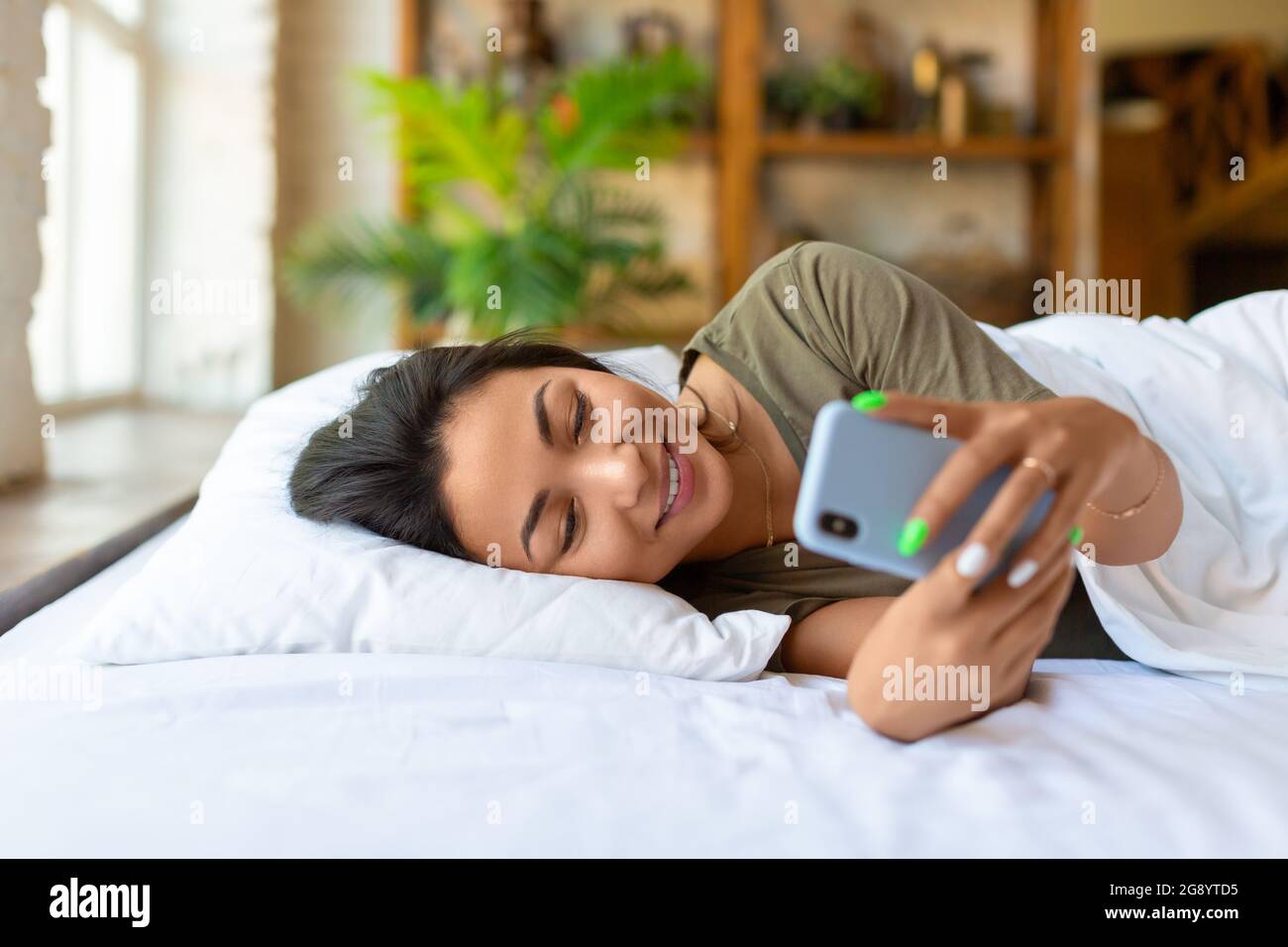 Une femme asiatique souriante est au lit avec un téléphone portable. Elle s'est réveillée et a vérifié les publications sur les médias sociaux. Concept de technologie moderne. Banque D'Images