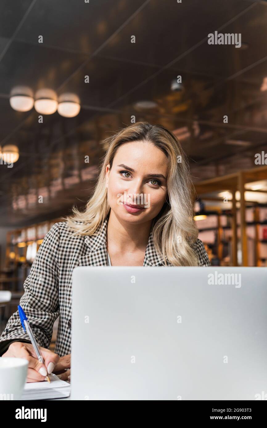 Une employée freelance écrit sur un journal tout en étant assise avec un ordinateur portable au café-restaurant Banque D'Images