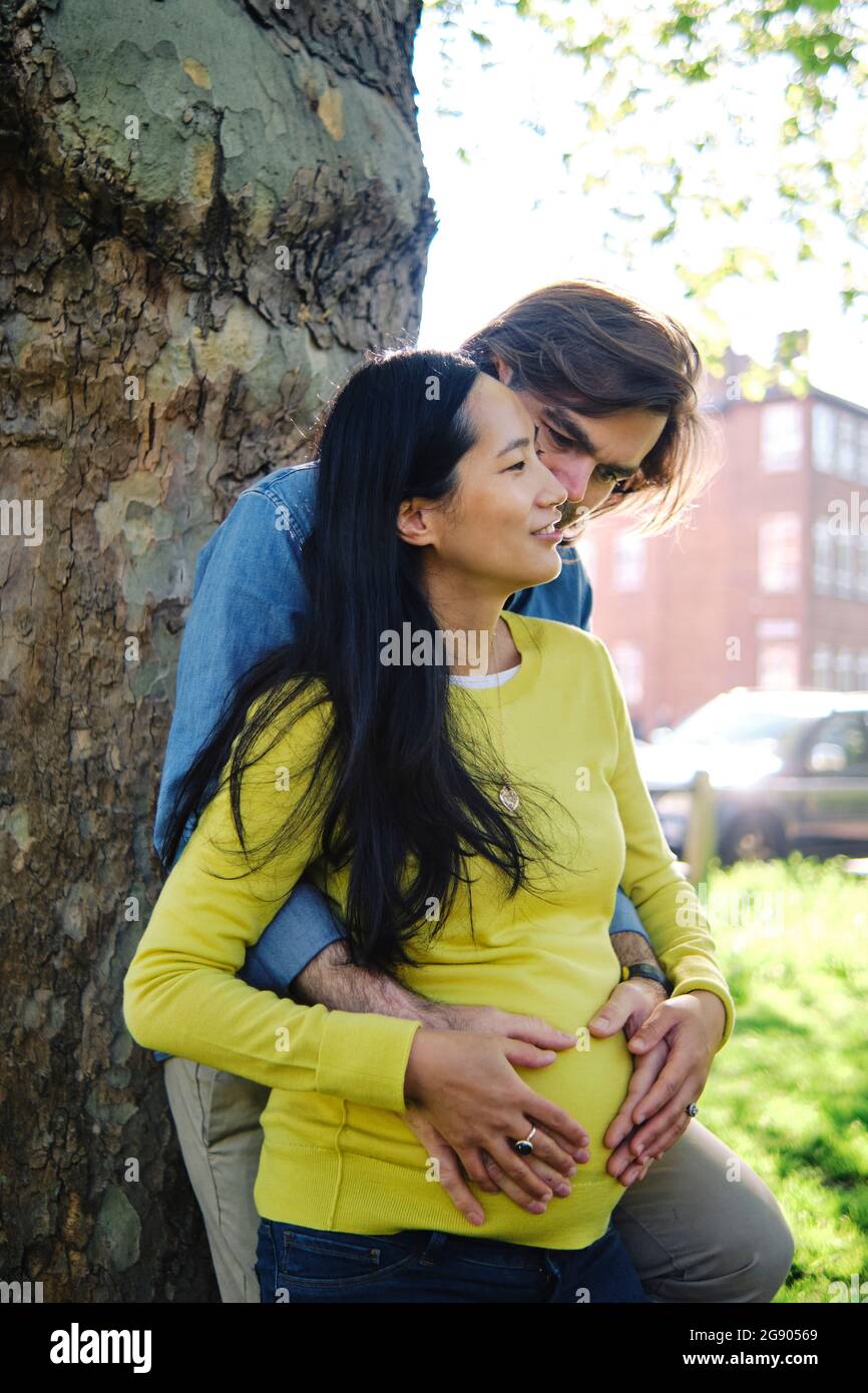 Homme embrassant la femme enceinte de derrière devant le tronc de l'arbre Banque D'Images