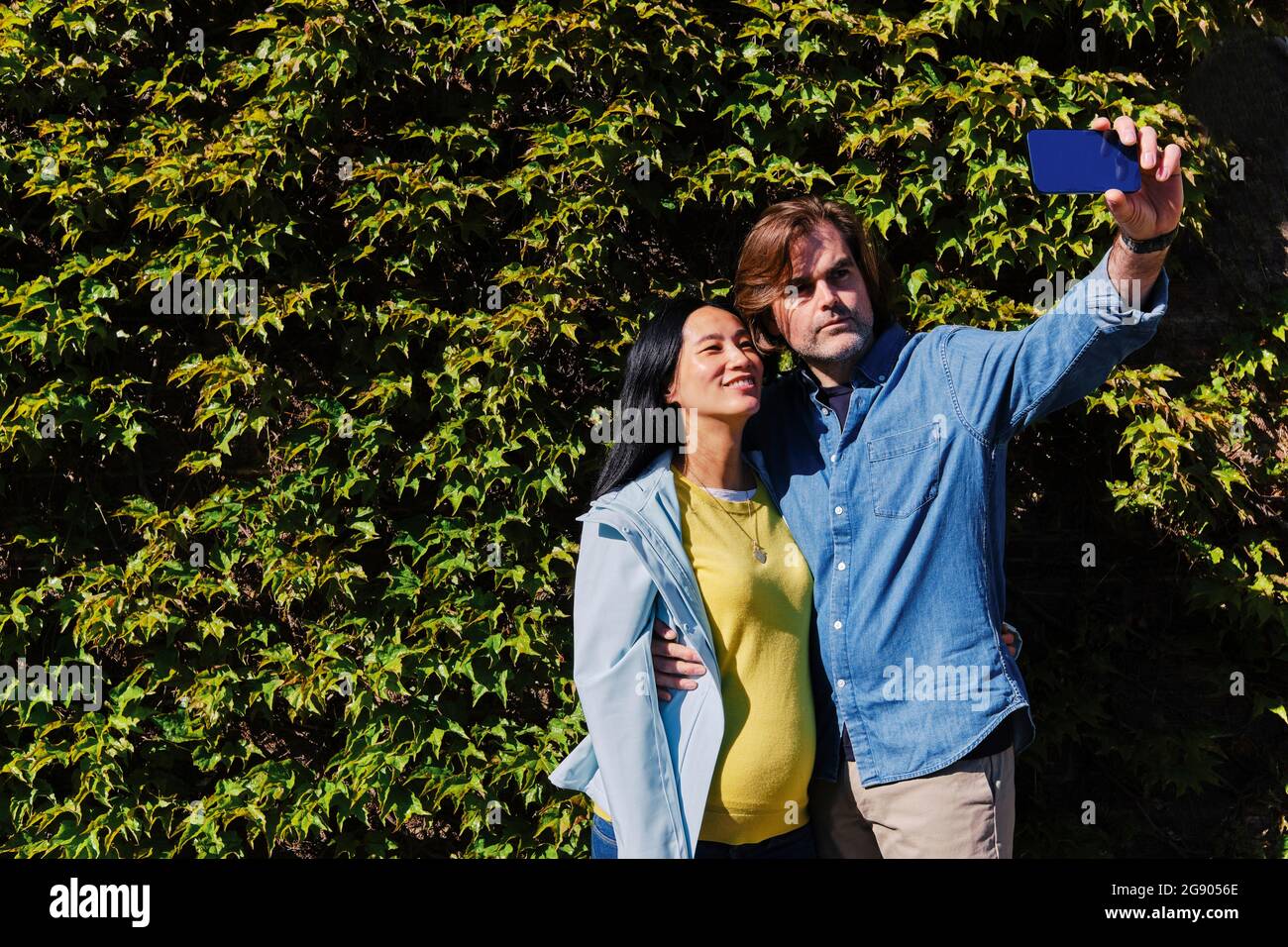 Homme prenant le selfie avec une femme enceinte à travers le smartphone devant la haie pendant la journée ensoleillée Banque D'Images