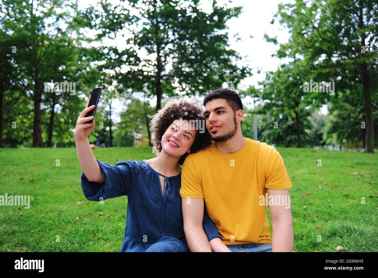 Jeune femme souriante emportant un selfie avec un petit ami à travers un smartphone tout en étant assise au parc Banque D'Images
