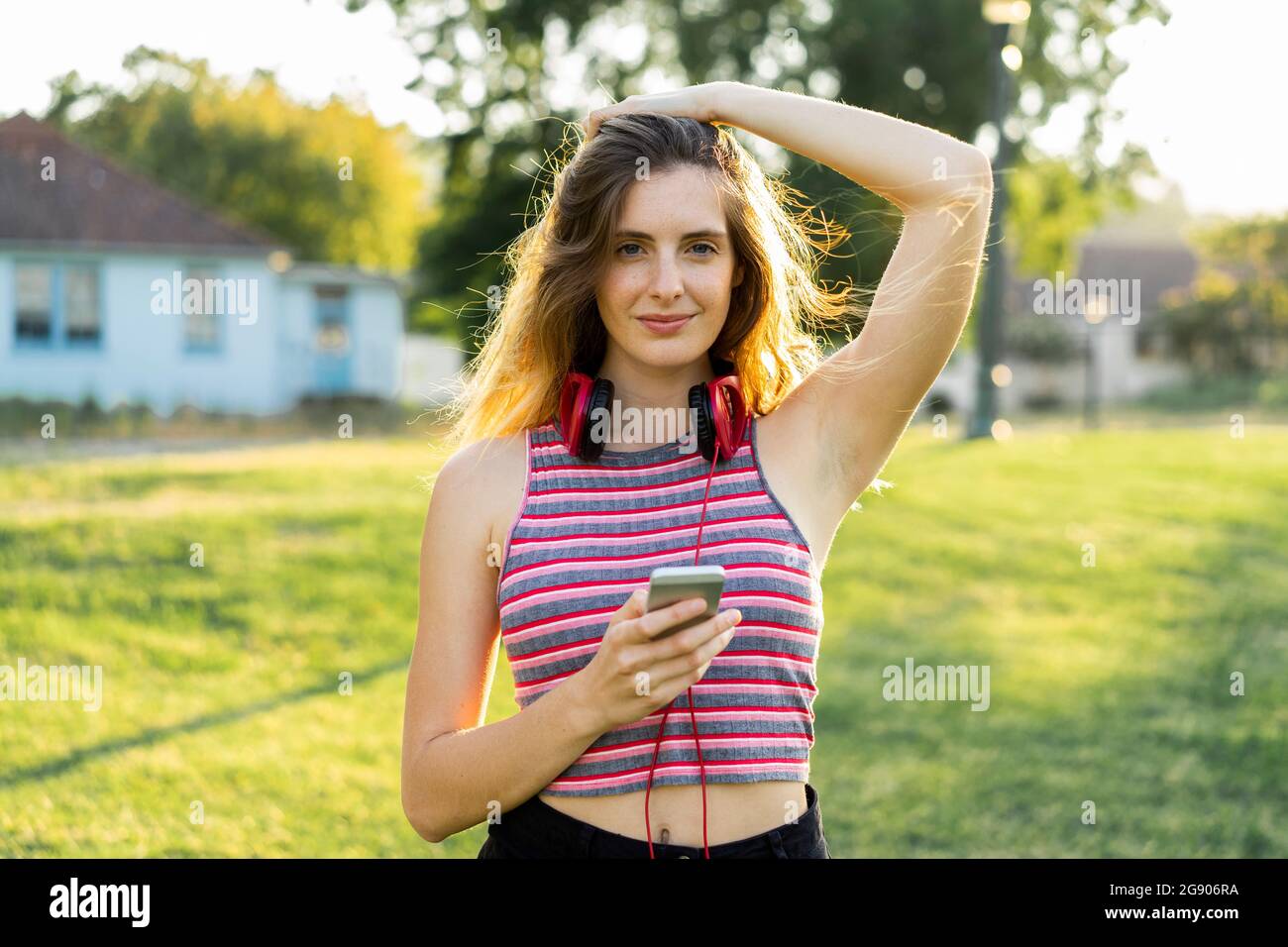 Belle jeune femme tenant un smartphone debout avec la main dans les cheveux au parc Banque D'Images