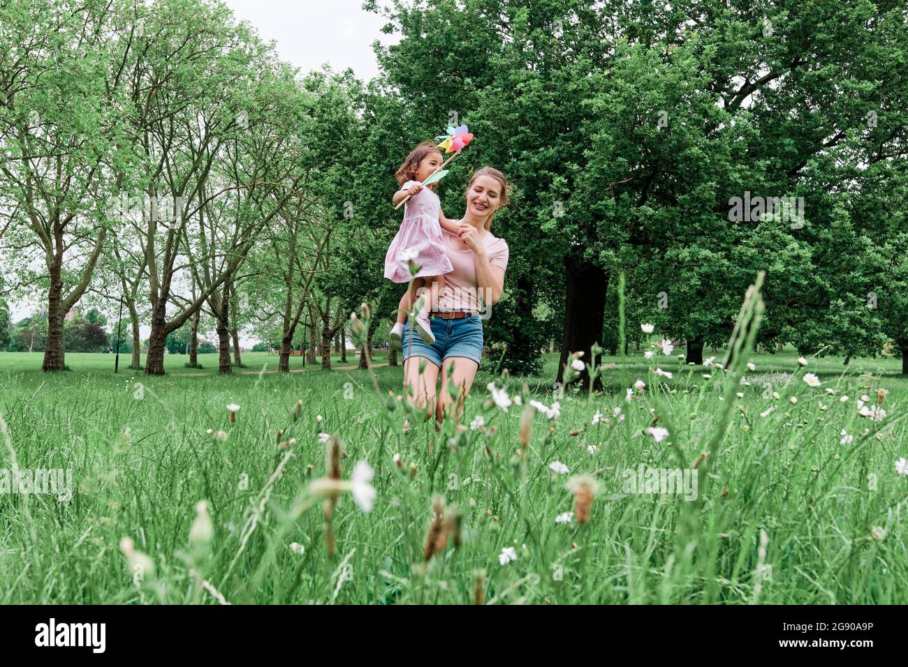 Bonne mère transportant sa fille avec un jouet à volant tout en marchant sur l'herbe au parc Banque D'Images