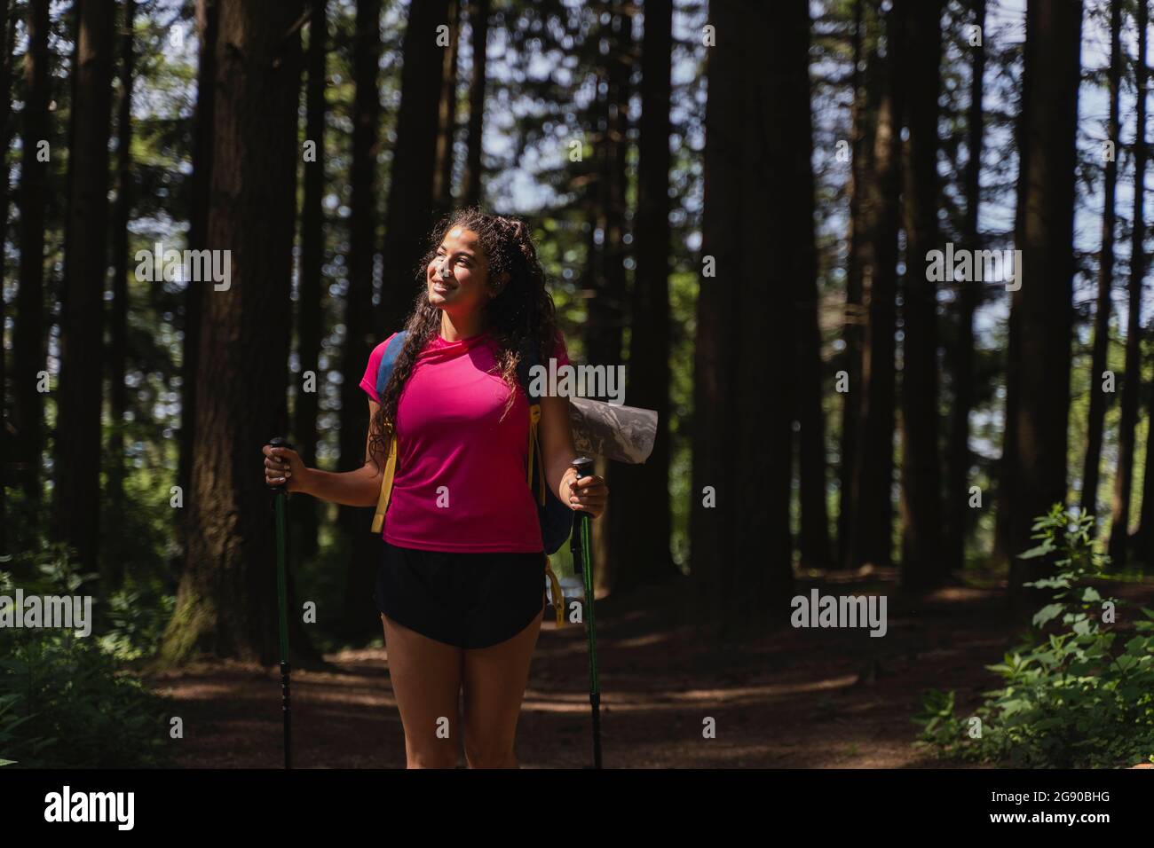 Une femme souriante qui regarde loin tout en marchant avec des bâtons de randonnée dans la forêt Banque D'Images