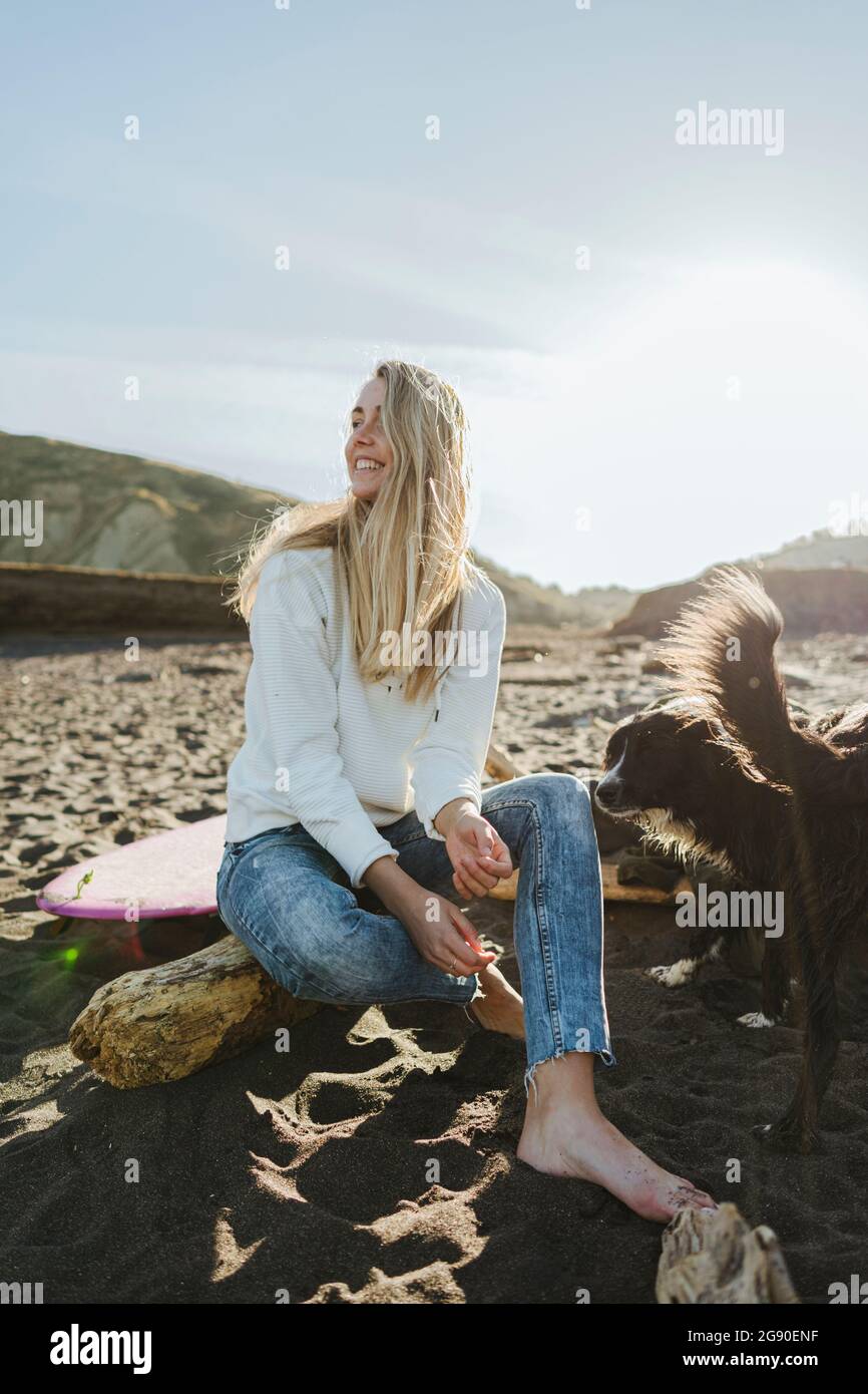 Femme blonde souriante qui regarde loin tout en étant assise près d'un chien à la plage Banque D'Images