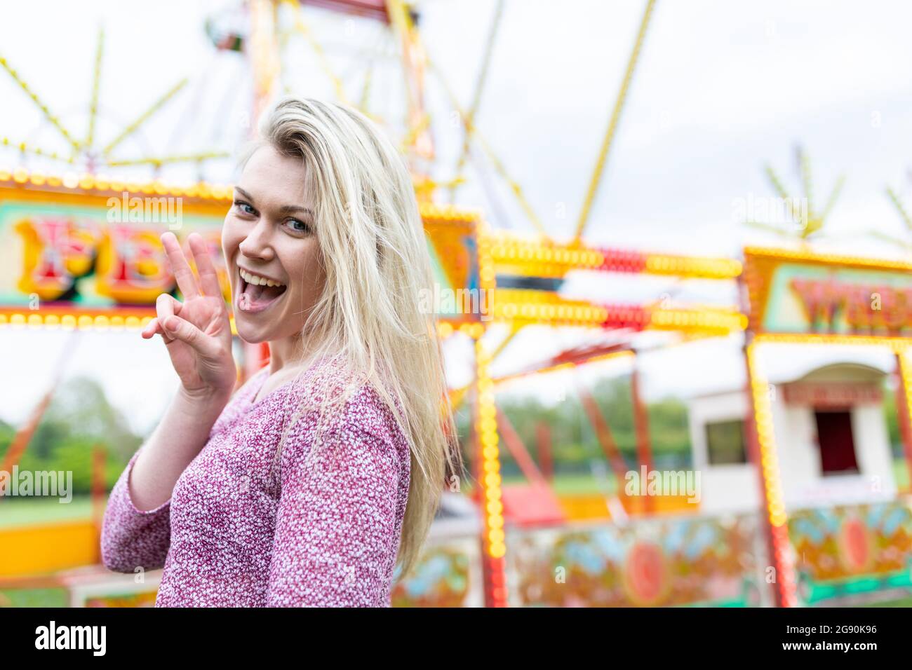 Une femme blonde gaie qui signe la paix tout en se tenant au parc d'attractions Banque D'Images