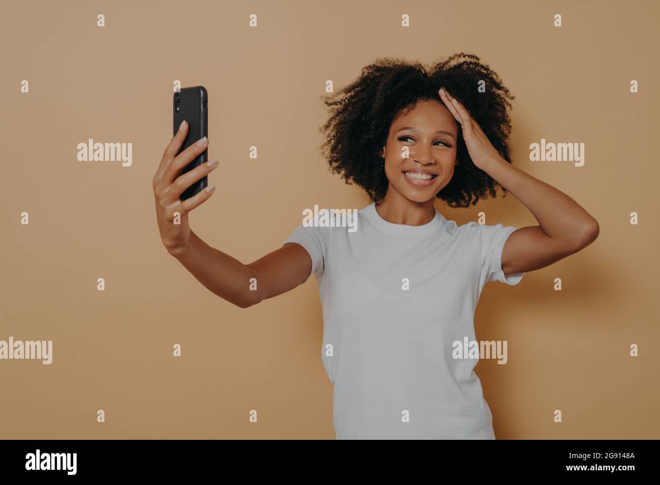 Bonne charmante femme à la peau sombre faisant un selfie photo sur un smartphone moderne contre un mur beige Banque D'Images