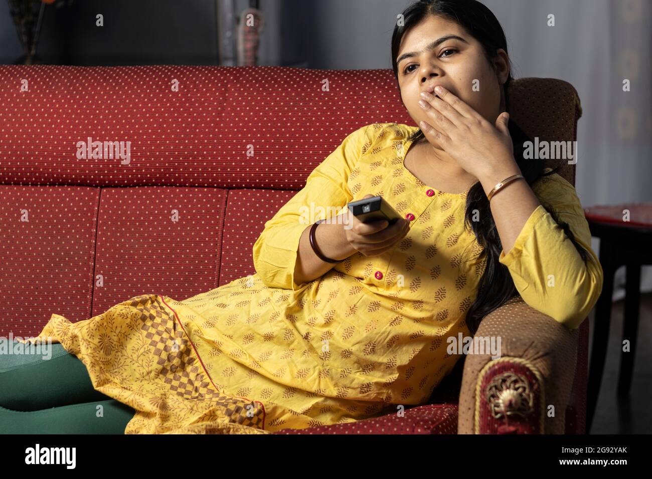 Une femme au foyer indienne bâillonnant tout en regardant la télévision tenant la télécommande dans la main, assis sur un canapé Banque D'Images