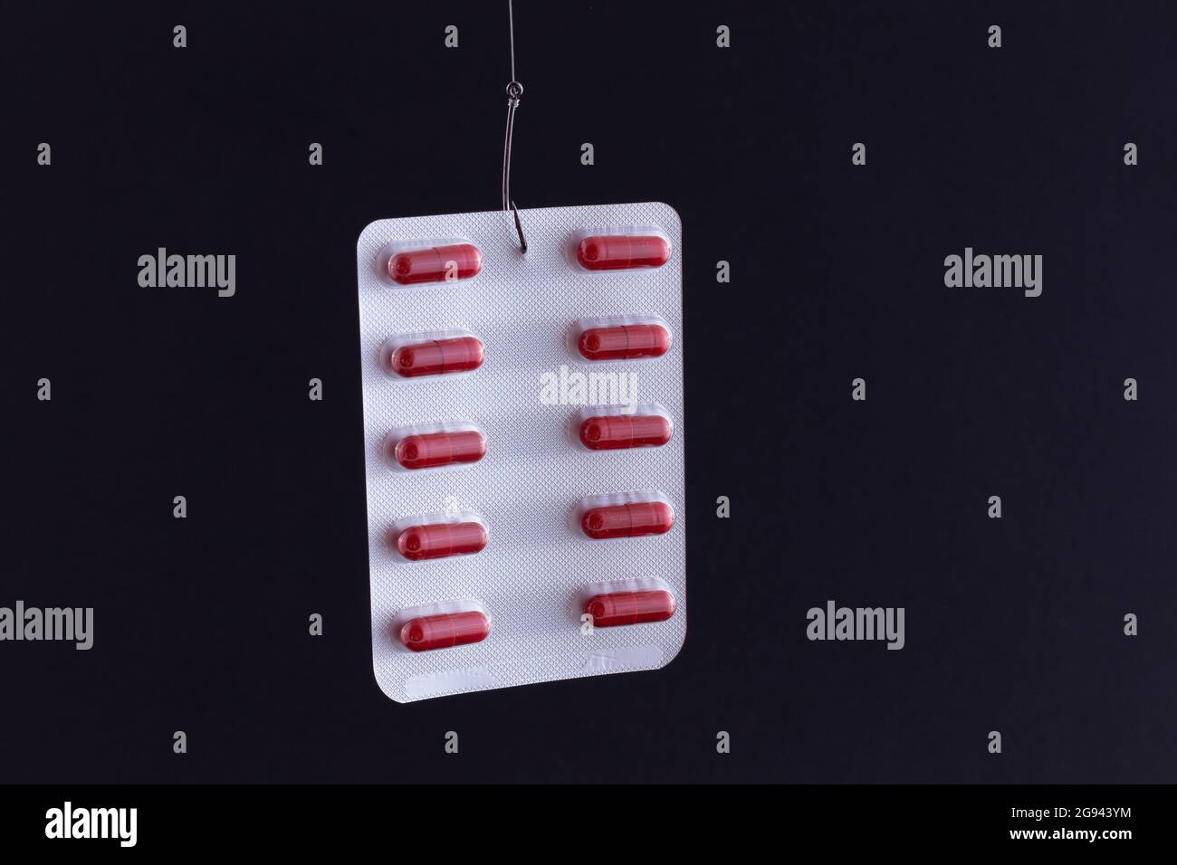 Le piège pharmaceutique et la toxicomanie - pilules rouges ou comprimés dans un emballage suspendu sur un crochet de pêche contre le fond noir Banque D'Images