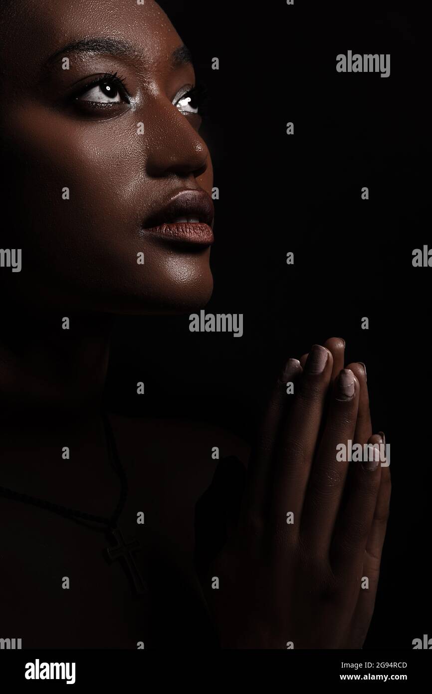 La femme prie. Portrait d'une belle femme africaine qui prie. Une jeune femme chrétienne est immergée dans la méditation tout en priant. Le visage et les mains sont éclairés. Banque D'Images