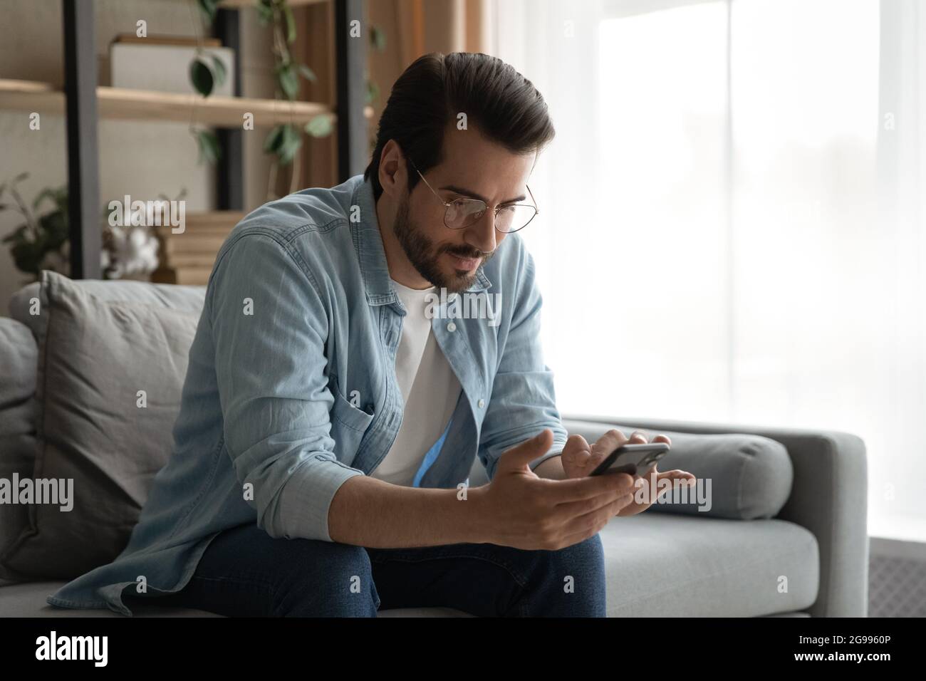 Un homme en lunettes s'assoit sur un canapé tenant le smartphone pour lire les messages Banque D'Images