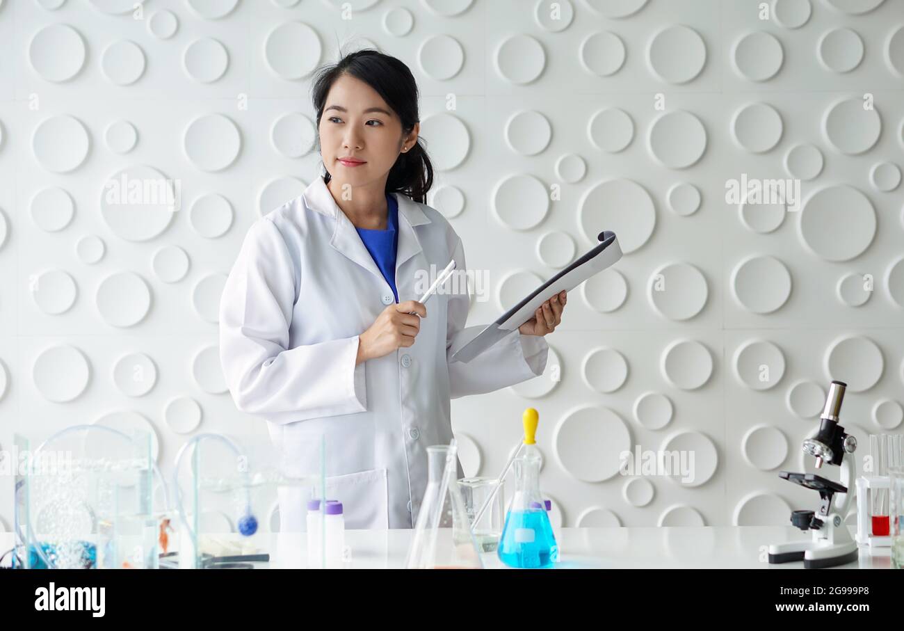 Jeune scientifique asiatique tenant un stylo et un presse-papiers. Elle regarda dehors tout en pensant à quelque chose. Atmosphère de travail en laboratoire chimique. Banque D'Images