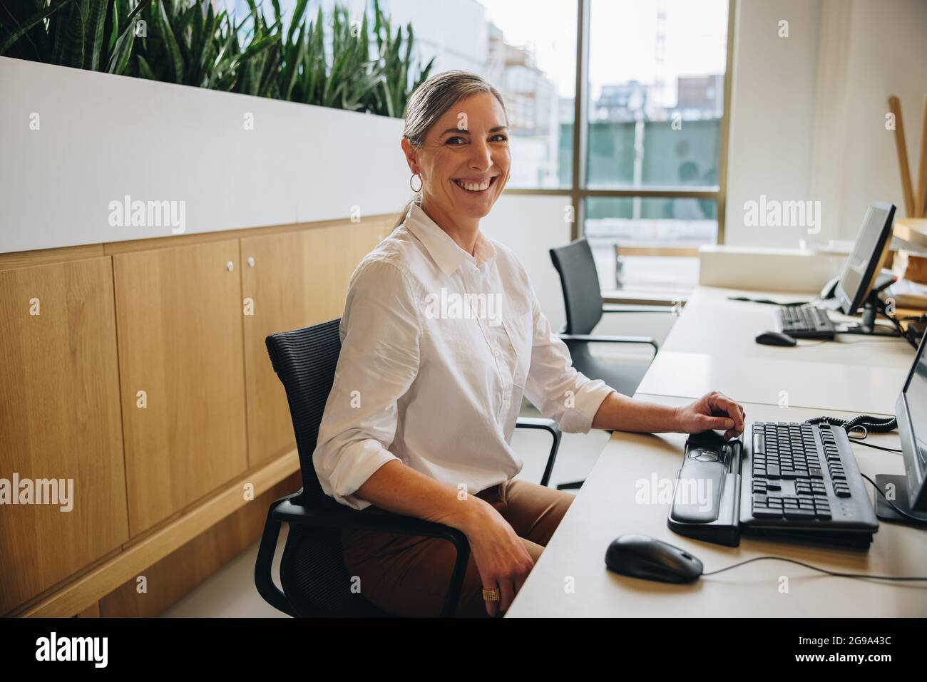Réceptionniste sympathique souriant à l'appareil photo tout en étant assis au bureau avec ordinateur dans le bureau. Une femme heureuse à la réception au travail. Banque D'Images
