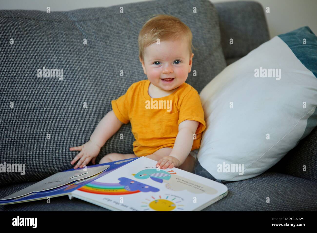 Un adorable enfant australien jouant avec un livre coloré sur un canapé dans une maison Banque D'Images