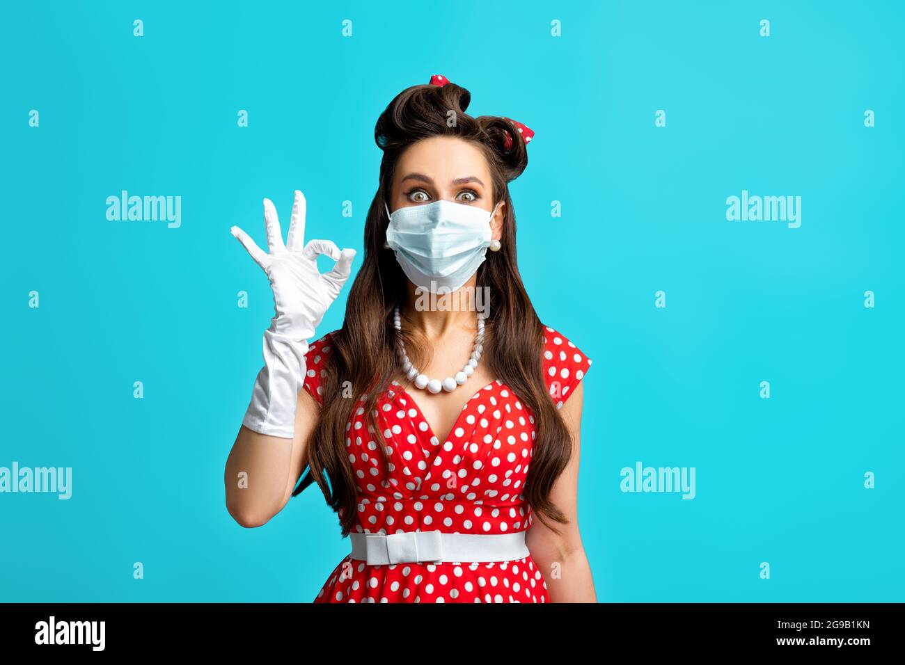 Portrait de la femme pinup en tenue rétro portant un masque pour éviter les covid, montrant un geste correct sur fond bleu Banque D'Images