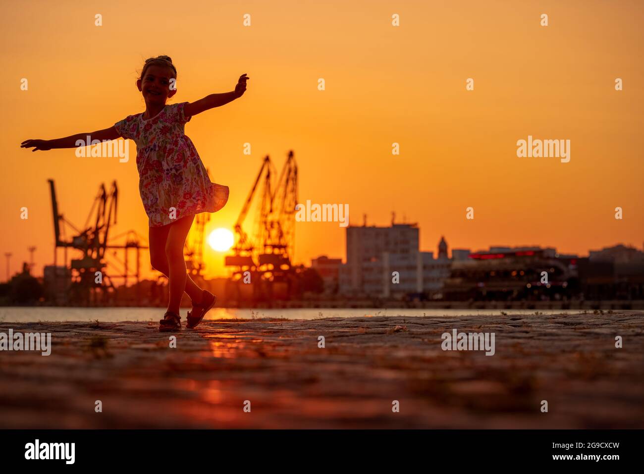 Petite fille dansant au coucher du soleil. Silhouttes de grues industrielles sur port de mer Varna, Bulgarie Banque D'Images