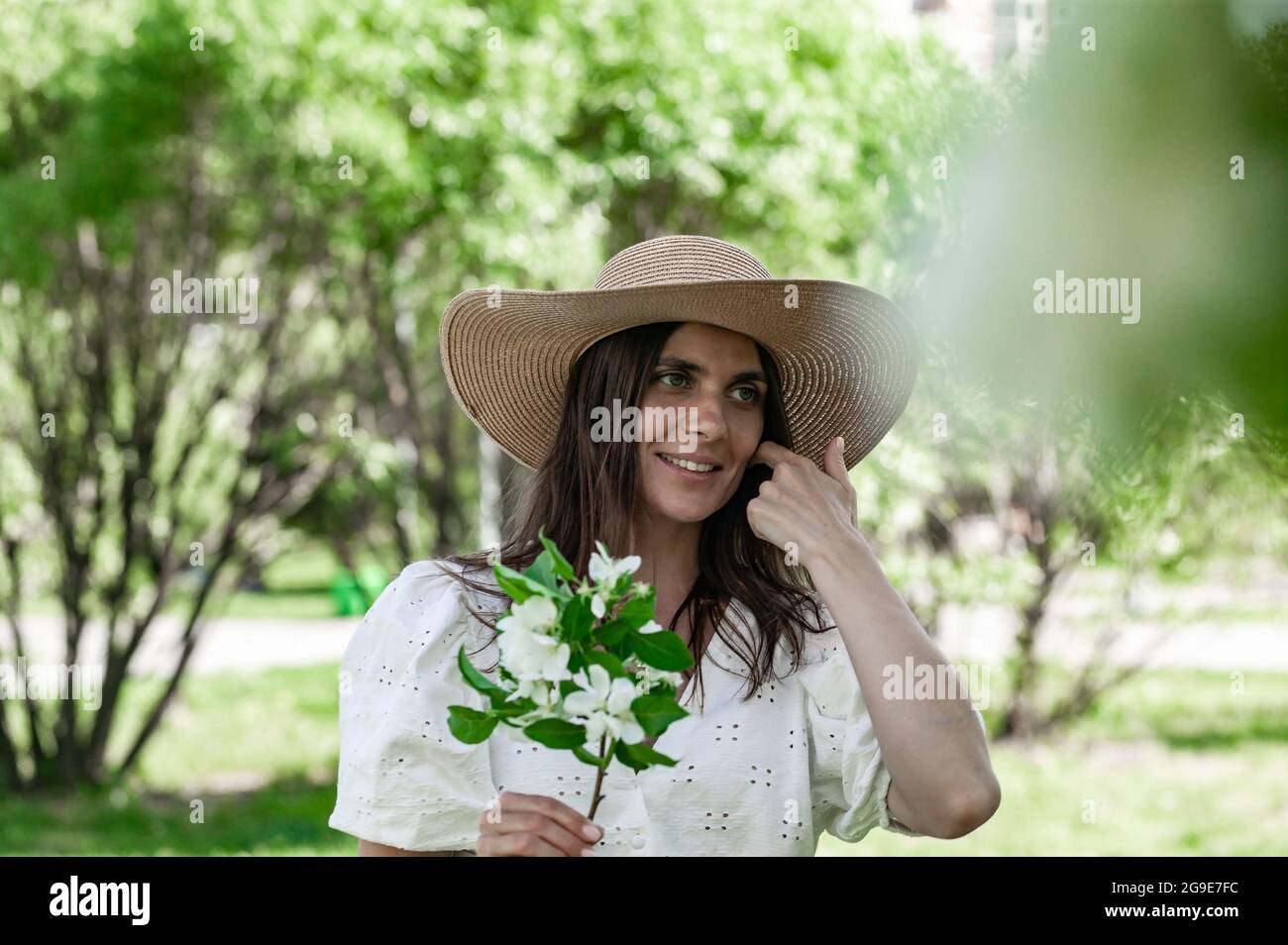 Jolie et attrayante femelle avec fleur de pomme. Jeune femme sur fond de parc de printemps. Mignonne souriante et heureuse jeune femme dans un chapeau de paille. Beau Banque D'Images