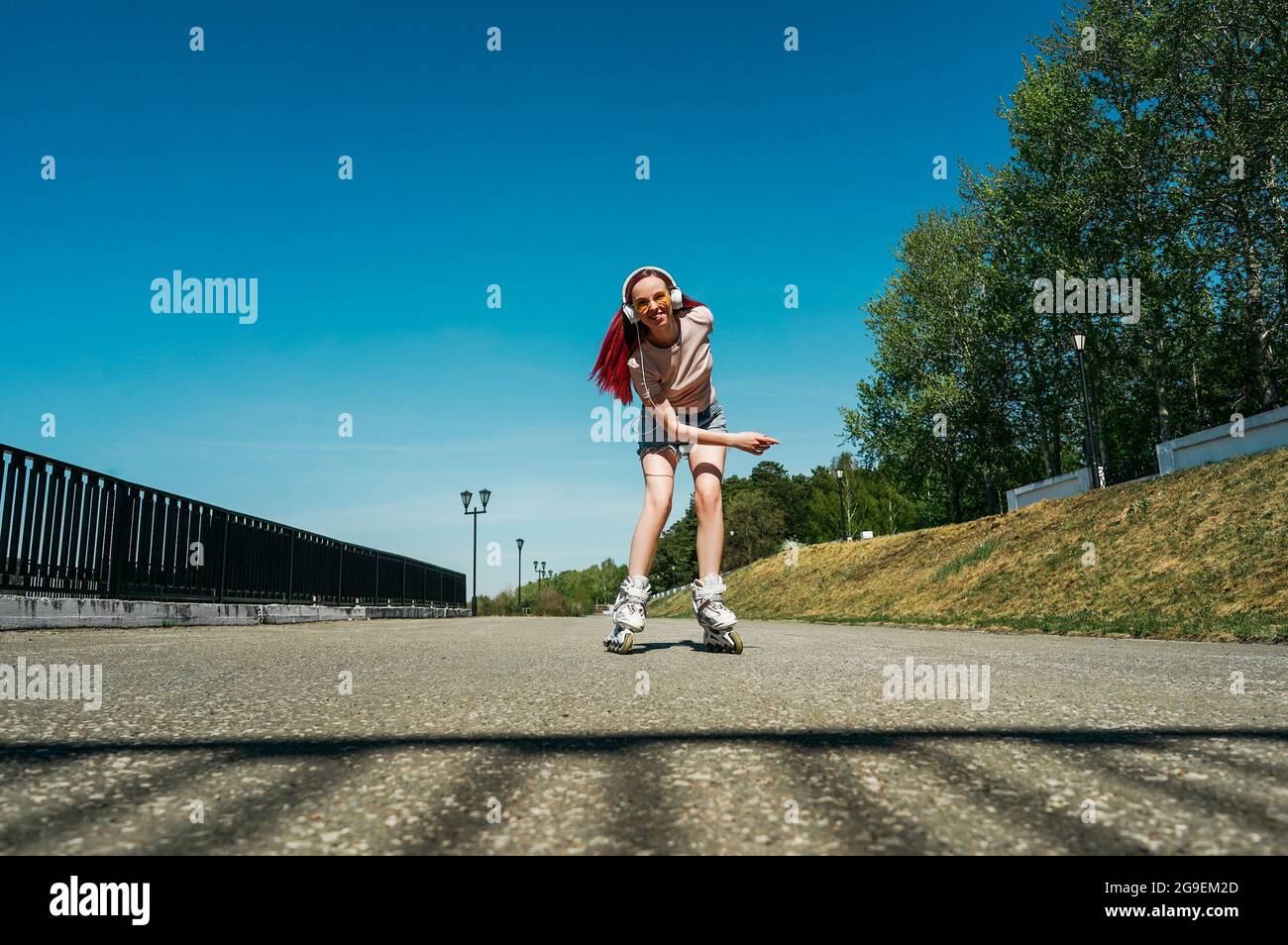 Une femme aux cheveux roses se déplace sur des patins à roulettes sur la route. Femme en lunettes de soleil jaunes et casque blanc. Banque D'Images