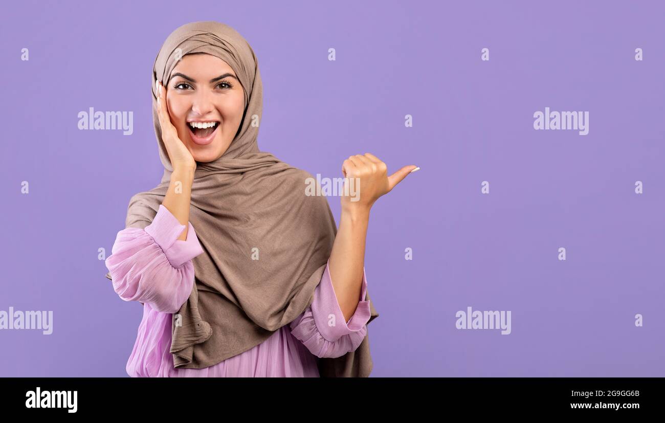 Ouah, vérifiez cela. Une dame arménienne musulmane excitée dans le hijab pointant vers l'espace libre sur fond violet, studio tourné, panorama. Surprise femme islamique dem Banque D'Images