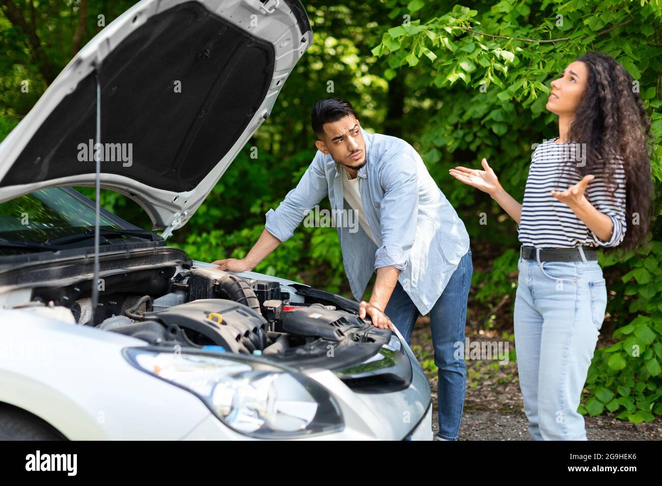 Un couple moyen-oriental contrarié ayant cassé une voiture pendant un voyage à la campagne, un homme arabe perplexe vérifiant le moteur, une femme brunette émotive regardant, triste mélangé Banque D'Images