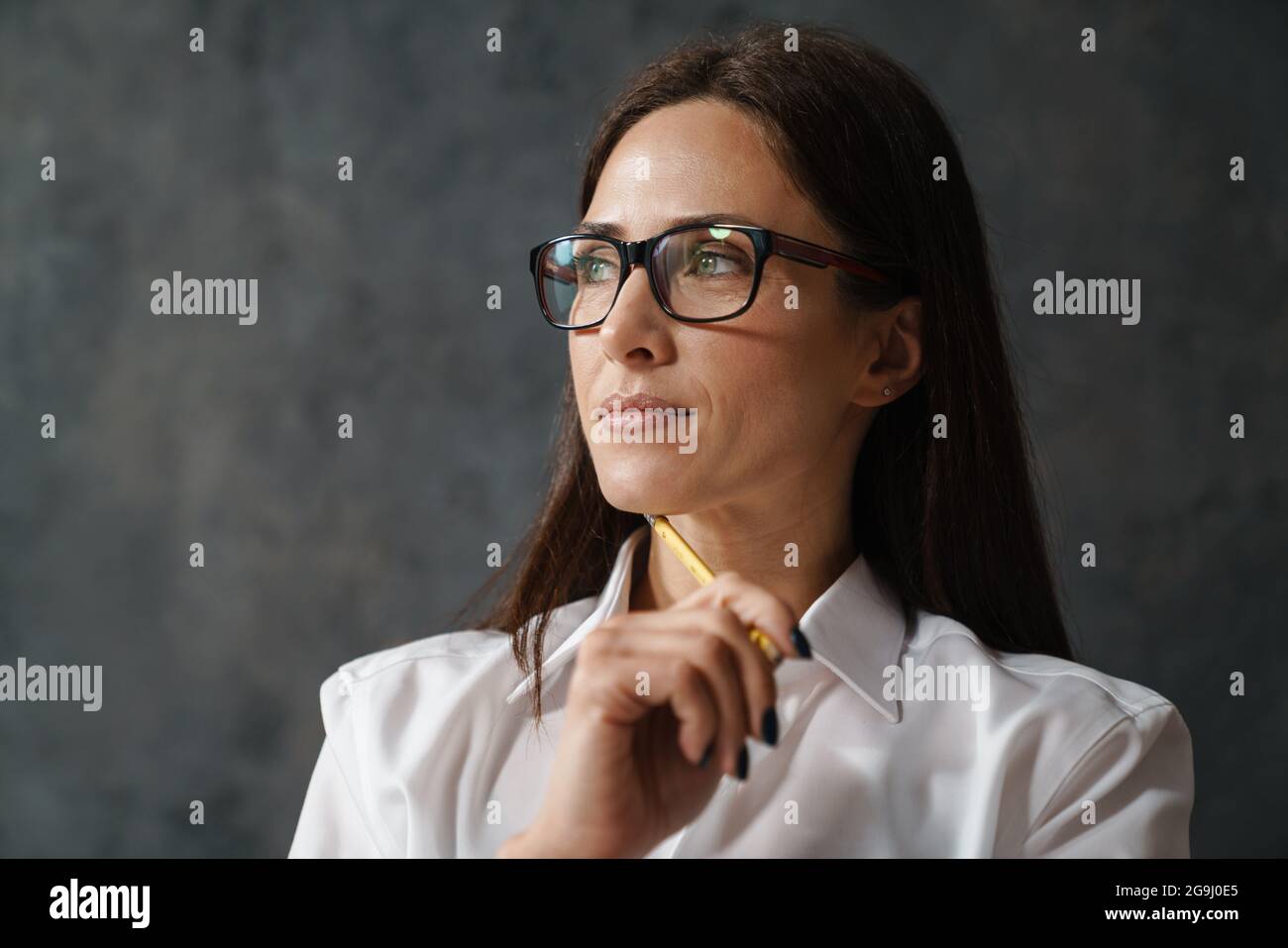 Femme de brunette pensive d'âge moyen en chemise blanche tenant un crayon sur son visage regardant de côté sur fond de mur sombre Banque D'Images