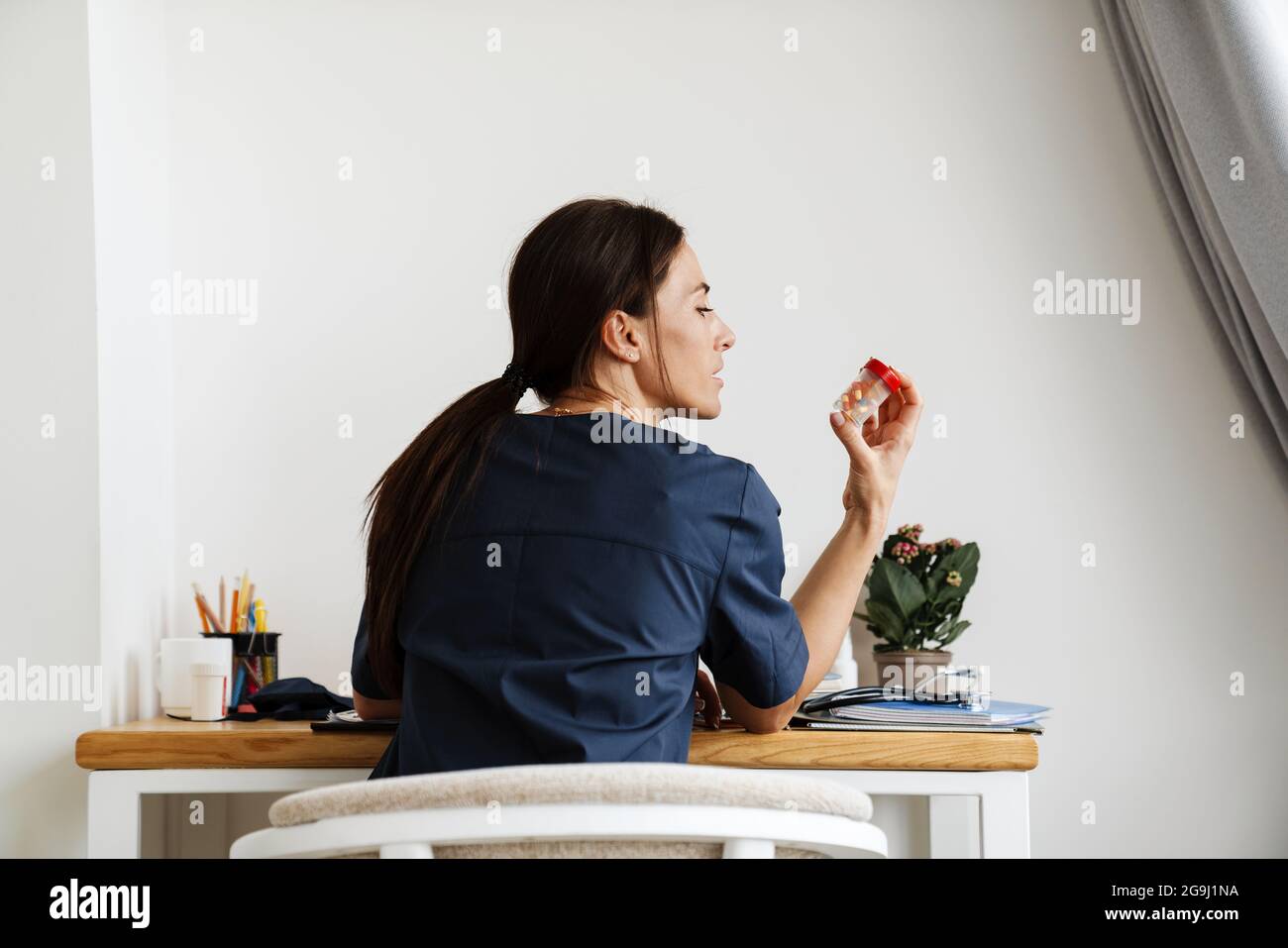 Vue arrière d'une femme médecin assise à une table et regardant latéralement un pot de pilules dans la chambre Banque D'Images