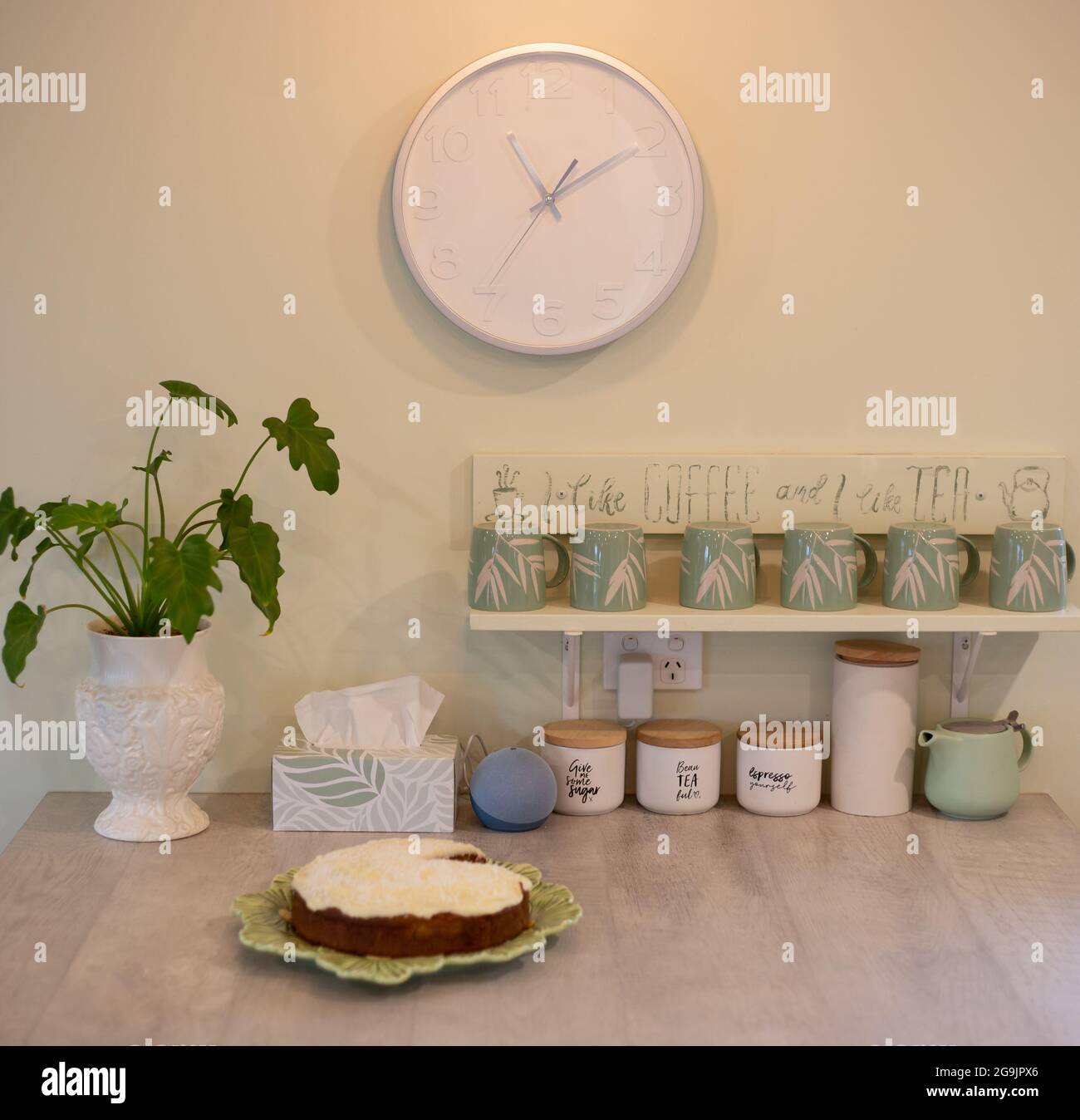 Cuisine banc d'îlot avec tasses à café, horloge, haut-parleur Alexa, plante maison et gâteau glacé Banque D'Images