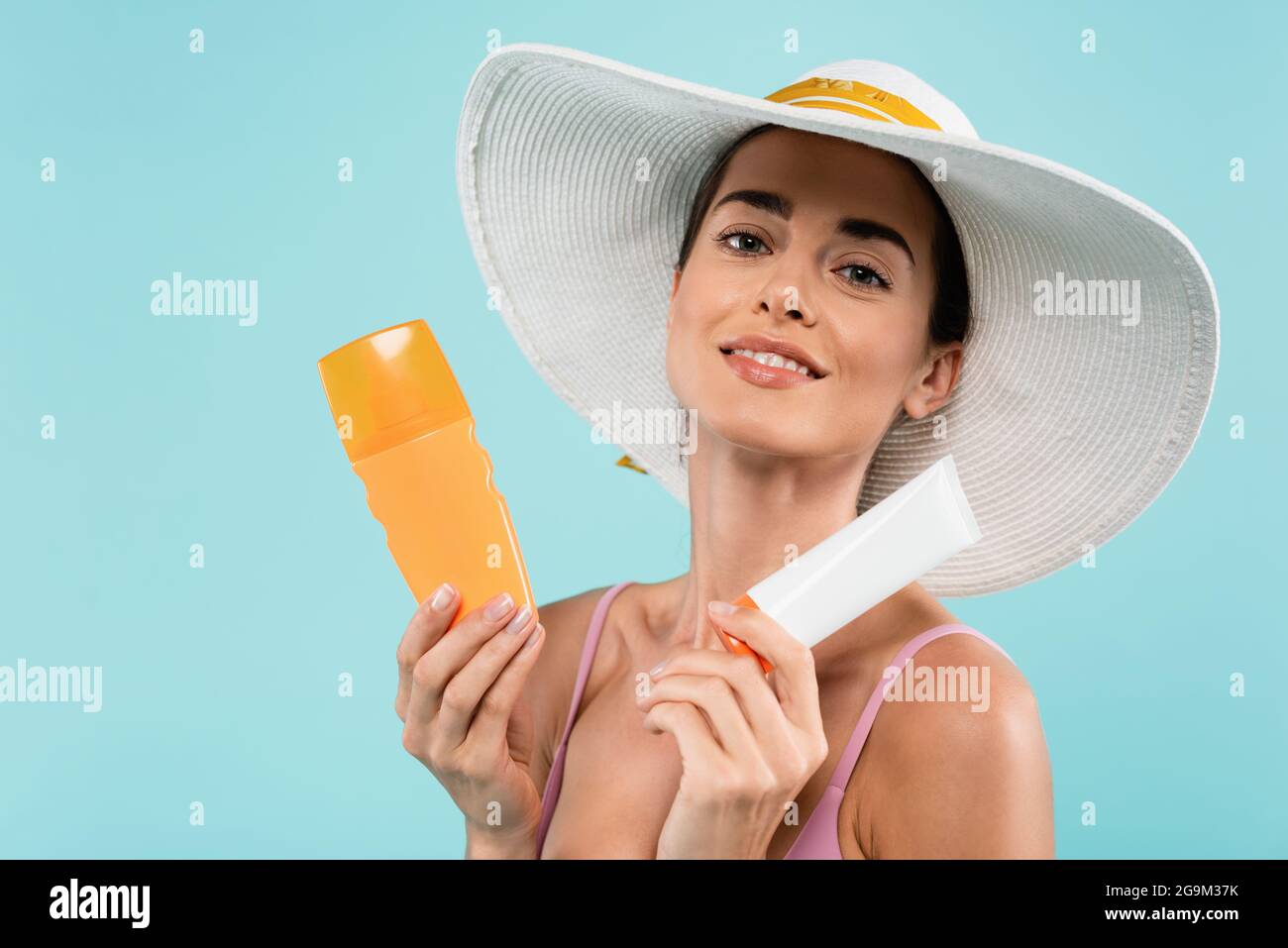 joyeuse femme en chapeau de soleil posant avec tube et bouteille de bloc de soleil isolé sur bleu Banque D'Images