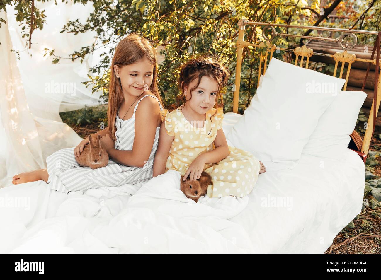 Petites jolies filles dans des robes d'été assis à l'extérieur avec des lapins, les enfants mignons regardant les lapins avec le sourire léger appréciant passer du temps avec Banque D'Images