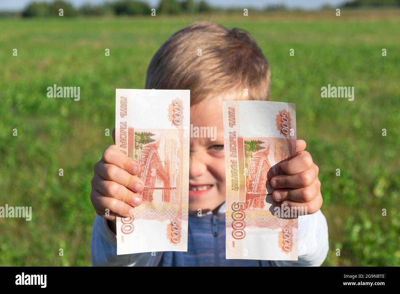 Un enfant satisfait, un garçon d'avant-garde, tient des roubles de papier dans ses mains à l'extérieur sur le fond d'un champ avec de l'herbe verte pendant un été ensoleillé Banque D'Images