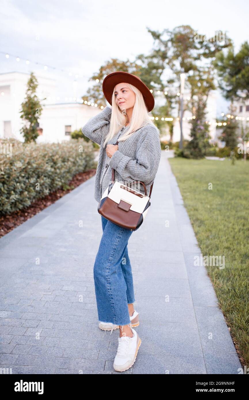 Sourire belle blonde adolescente 17-18 ans porter chapeau élégant, sac en  cuir et pull gris tricoté marcher dans la rue de la ville dans le parc à  l'extérieur. Réglage Photo Stock -