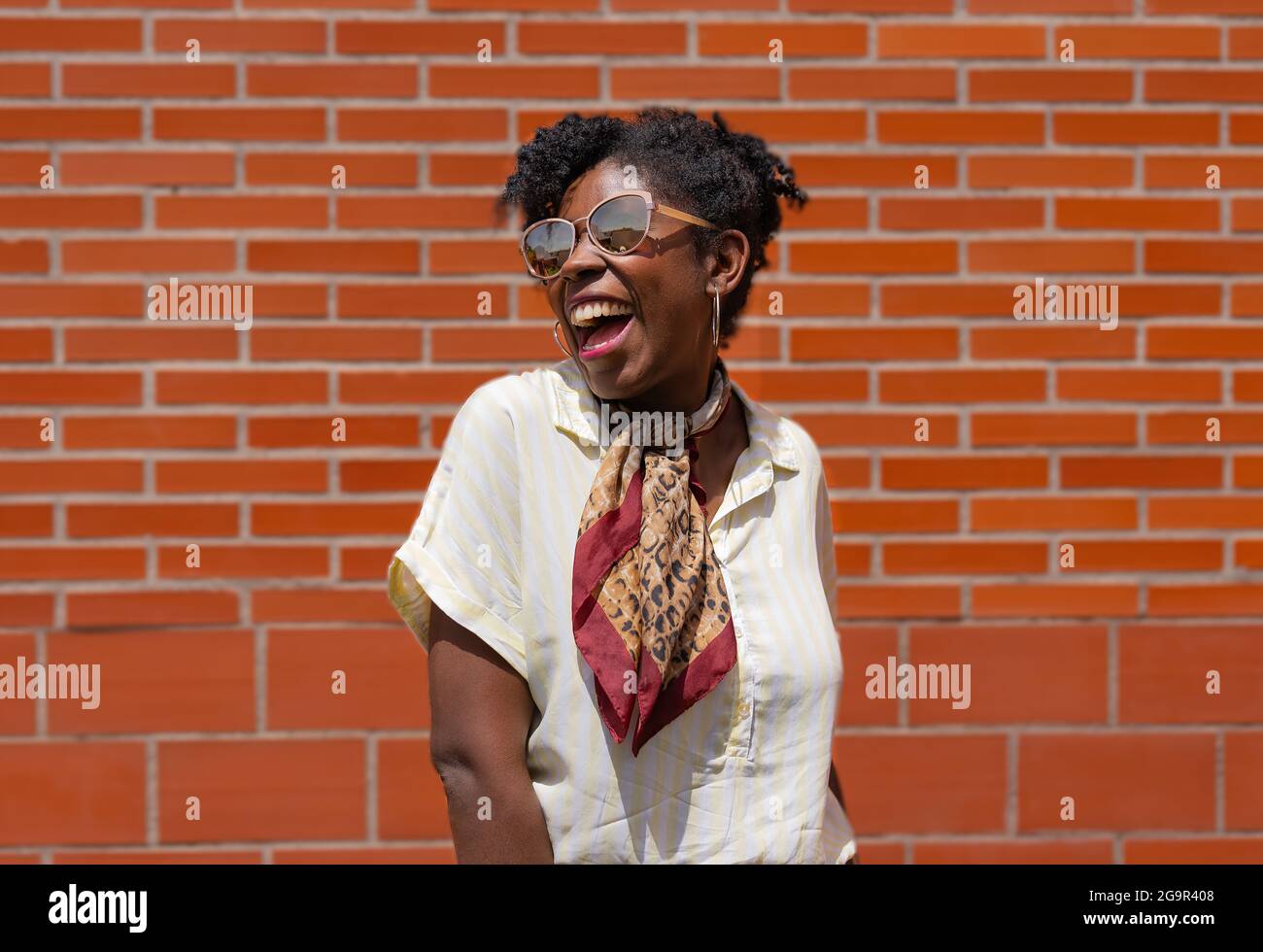 Femme noire ravie dans des vêtements élégants et des lunettes de soleil souriant et regardant loin tout en se tenant contre le mur de brique dans la rue Banque D'Images