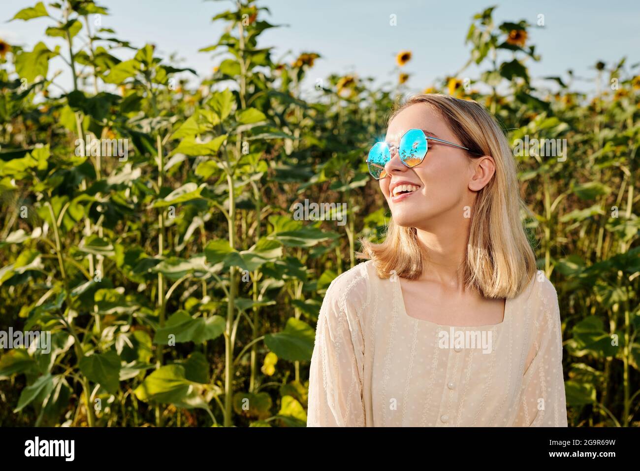 Joyeuse jeune femme blonde en lunettes de soleil et robe blanche debout contre le champ de tournesol devant la caméra avec ciel bleu en arrière-plan Banque D'Images