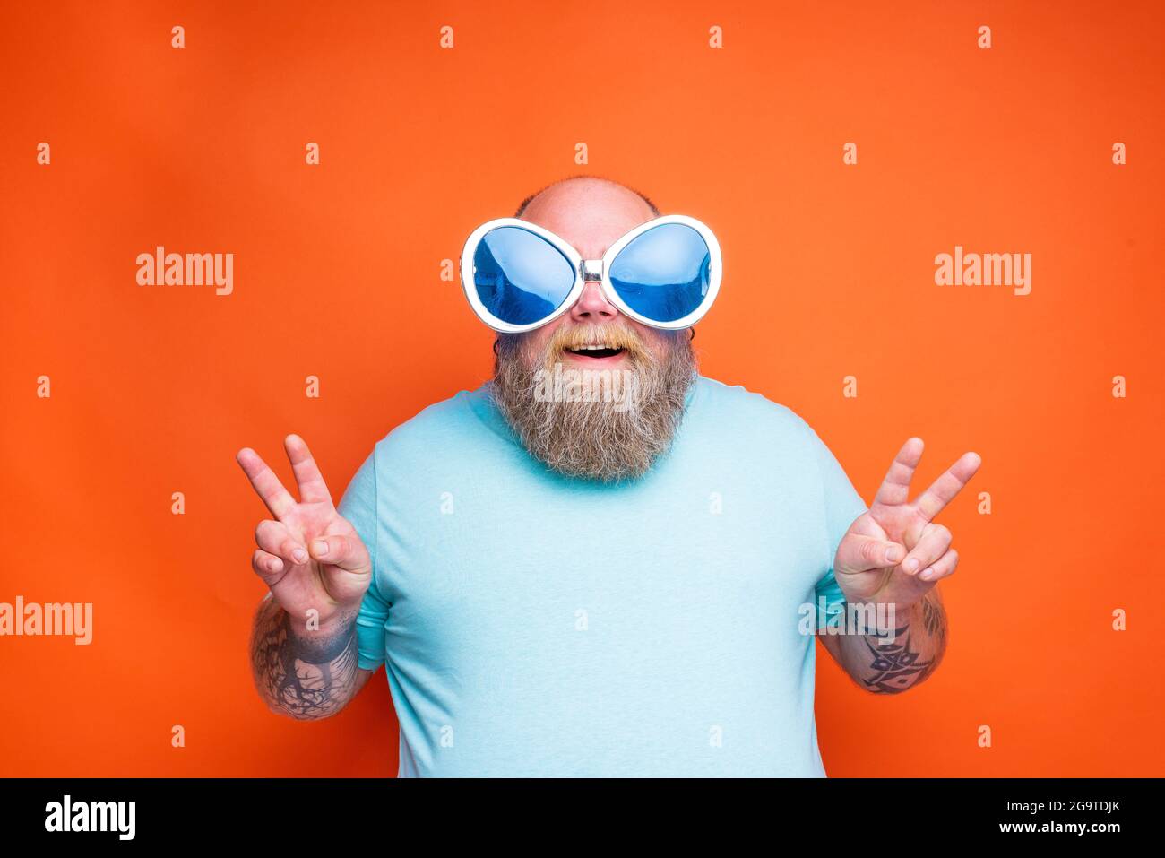 Gros Happy man avec la barbe, les tatouages et les lunettes de soleil fait le signe de la victoire avec ses mains Banque D'Images