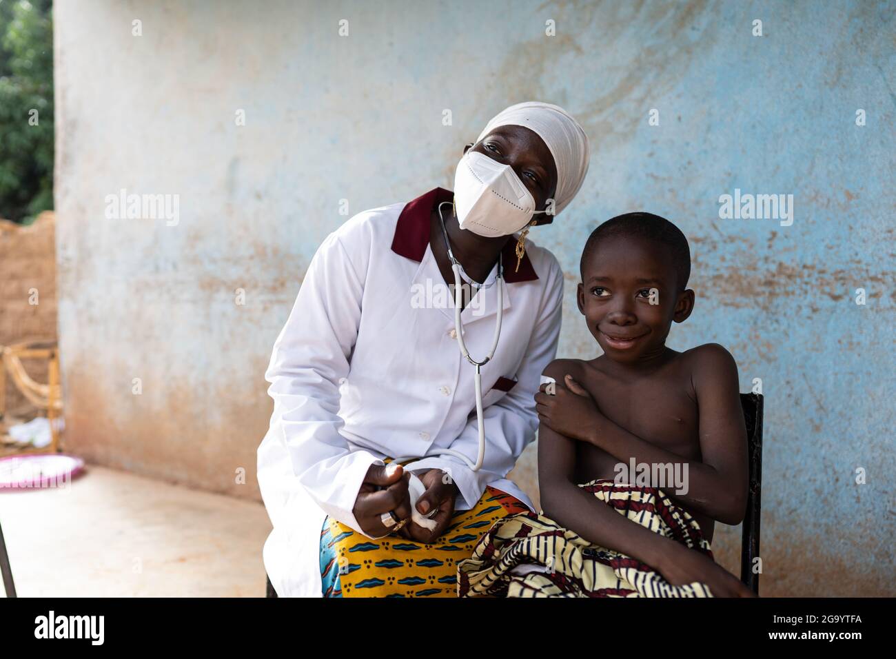 Infirmière noire avec masque facial et stéthoscope et petit enfant africain sans chirerie assis à côté d'elle à l'extérieur d'un petit dispensaire de village africain, les deux s Banque D'Images