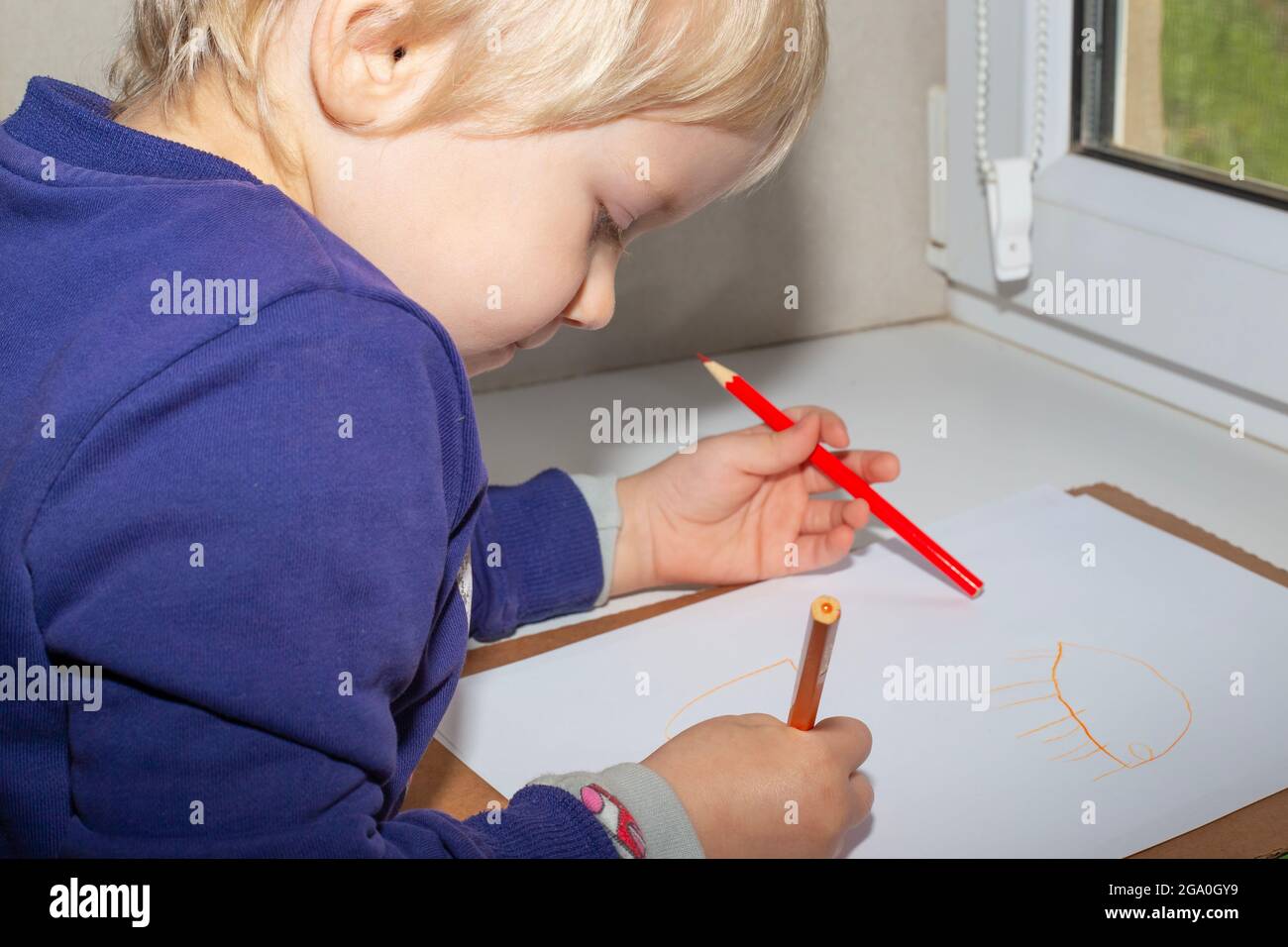 Un petit garçon aux cheveux justes avec des crayons dans ses mains tire sur une feuille blanche sur le rebord de la fenêtre. Développement de la petite enfance. Banque D'Images