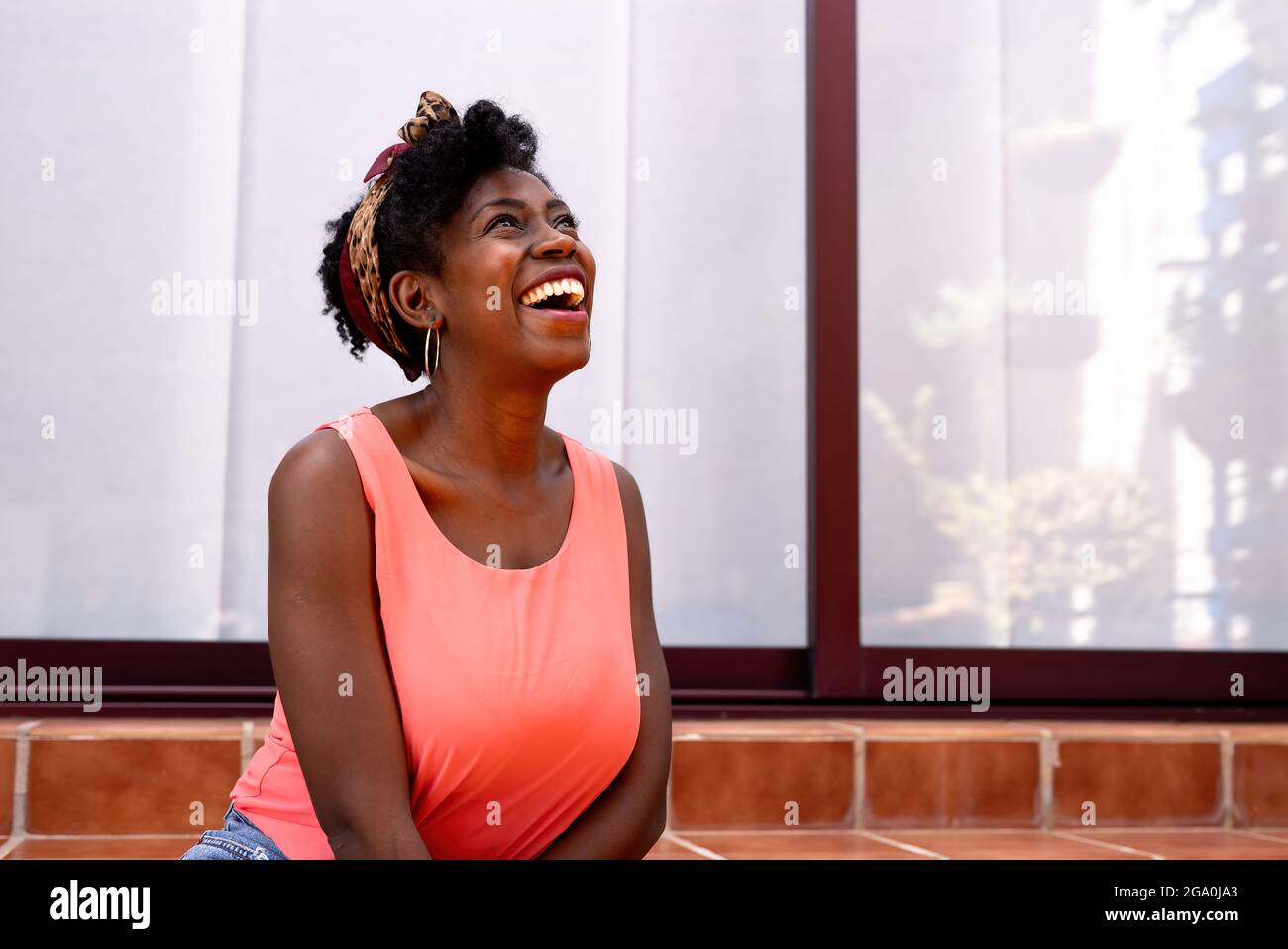 Jeune femme afro-américaine joyeuse en haut rose décontracté et serre-tête riant joyeusement tout en s'amusant près d'un bâtiment moderne avec des murs en verre Banque D'Images