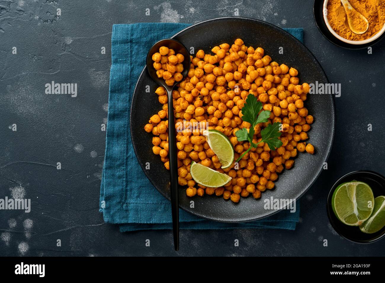 Pois chiches frits au curcuma avec persil et citron vert dans une assiette noire sur un fond de table noir ancien. Pois chiches épicés grillés ou chana ou chole indien, Banque D'Images