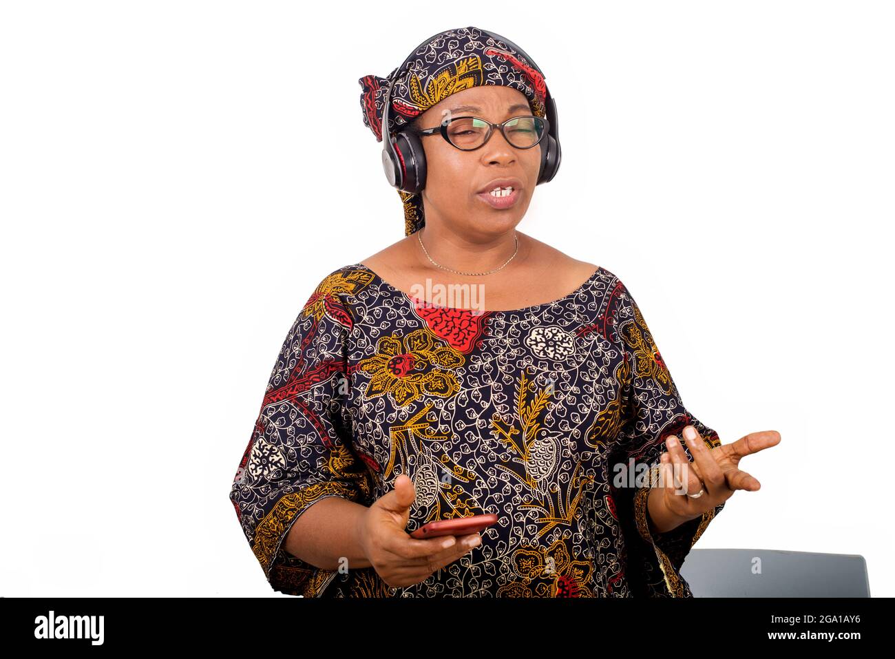 femme africaine mûre en tissu de longe, parlant et regardant surpris tout en ayant le geste de main tout en écoutant de la musique avec des écouteurs. Banque D'Images