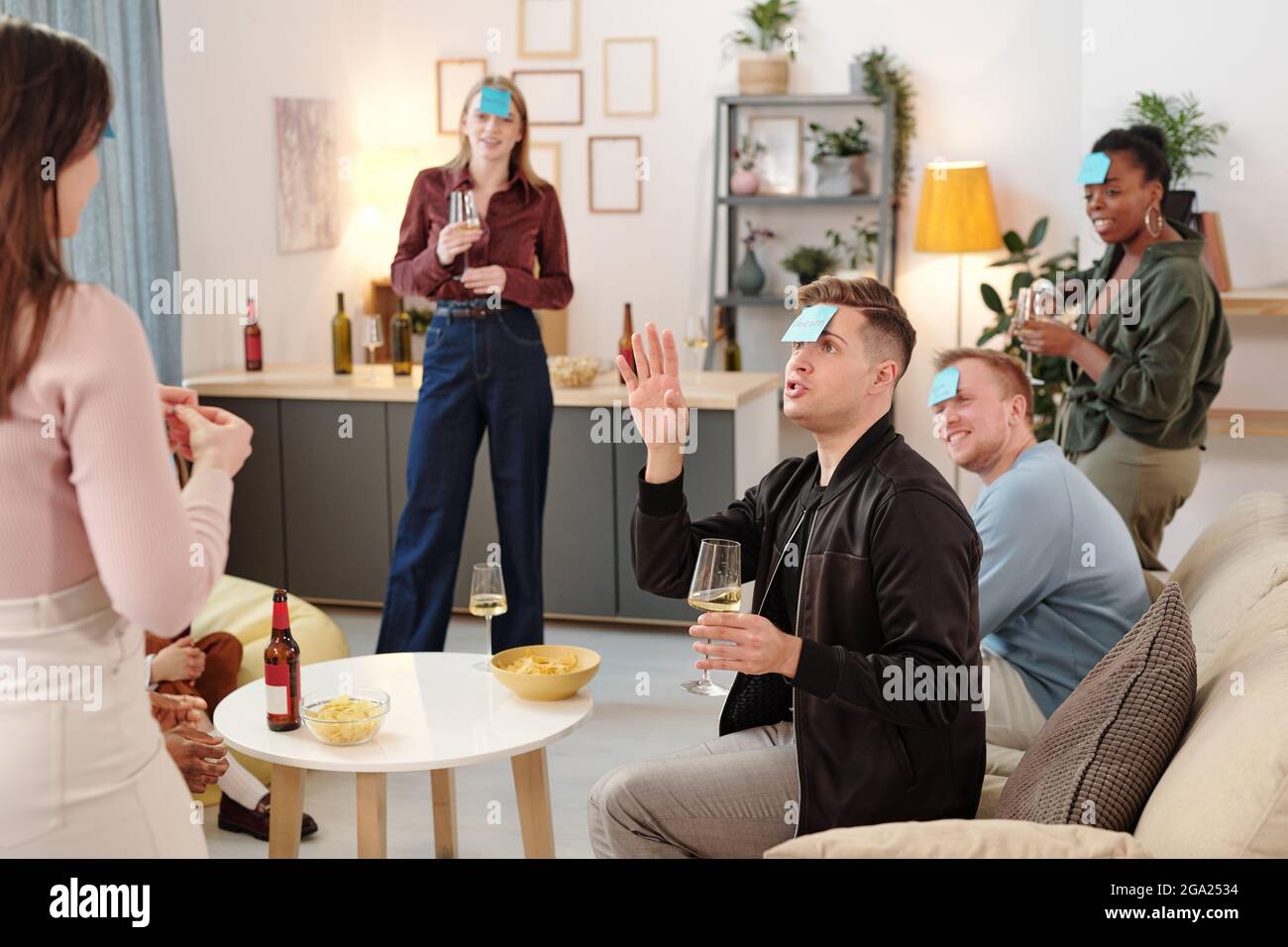Un groupe de jeunes amis interculturels joyeux en vêtements décontractés intelligents se sont rassemblés dans un salon et dans un jeu de noms, en riant Guy devinant qui il est Banque D'Images