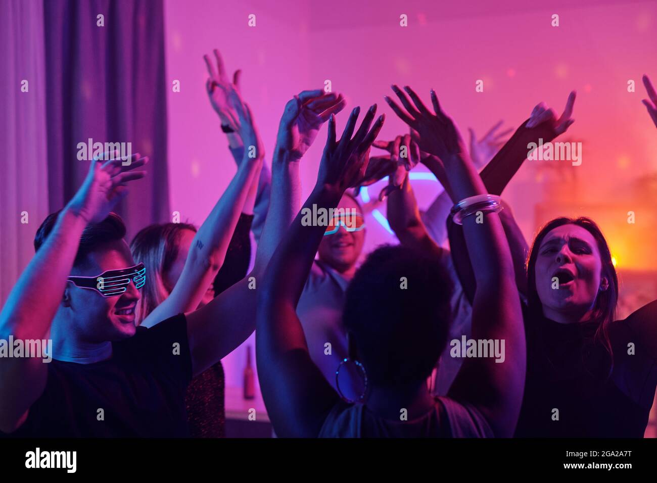 De jeunes amis glamour et interculturels élèvent leurs bras tout en dansant ensemble à la maison, dans la salle de séjour éclairé par un éclairage rose Banque D'Images