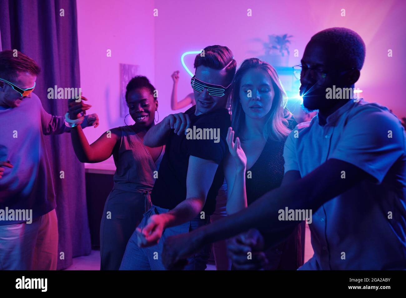 Groupe de jeunes amis interculturels joyeux dans la danse chic de vêtements décontractés excithly à la fête à la maison dans la salle de séjour éclairé avec des lumières roses Banque D'Images