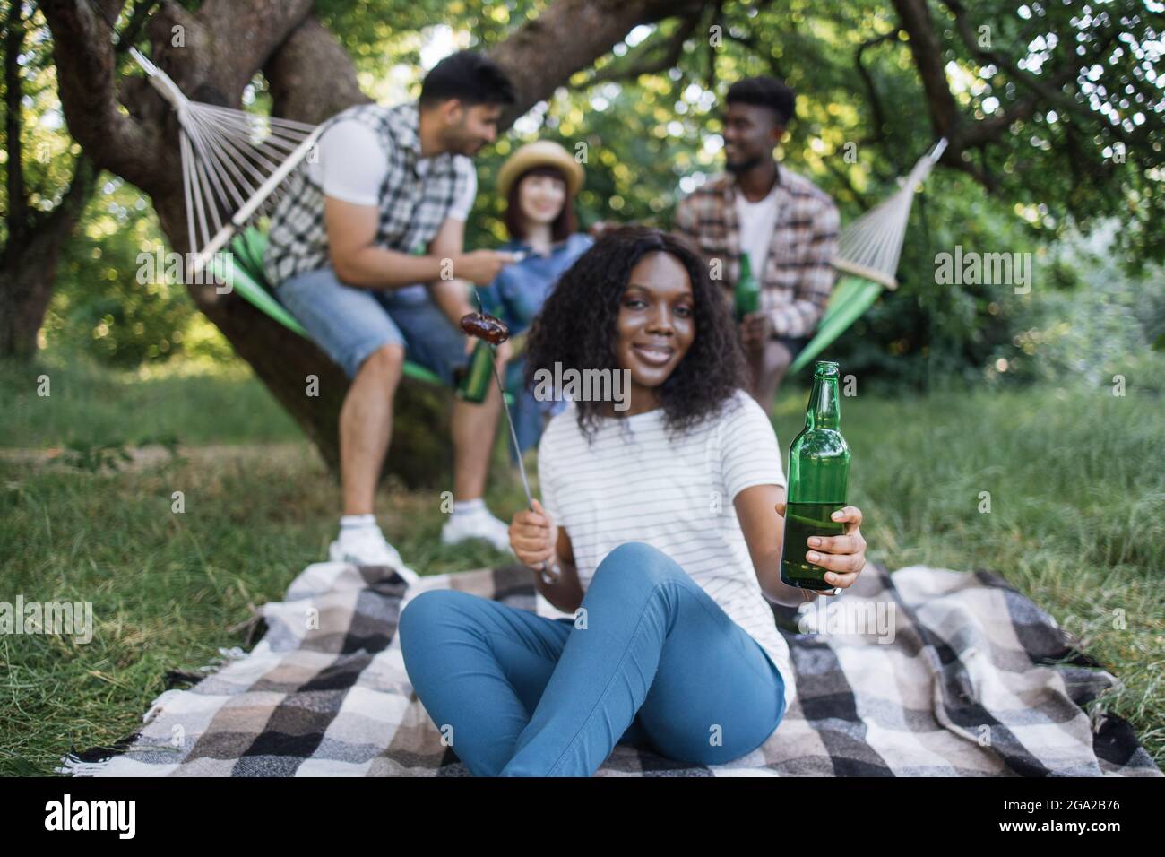 Femme afro americaine dans une tenue decontractee posant au jardin avec des saucisses grillees et de la biere froide. Jolie dame pique nique avec trois amis a l exterieur. Temps de relaxation Photo St...